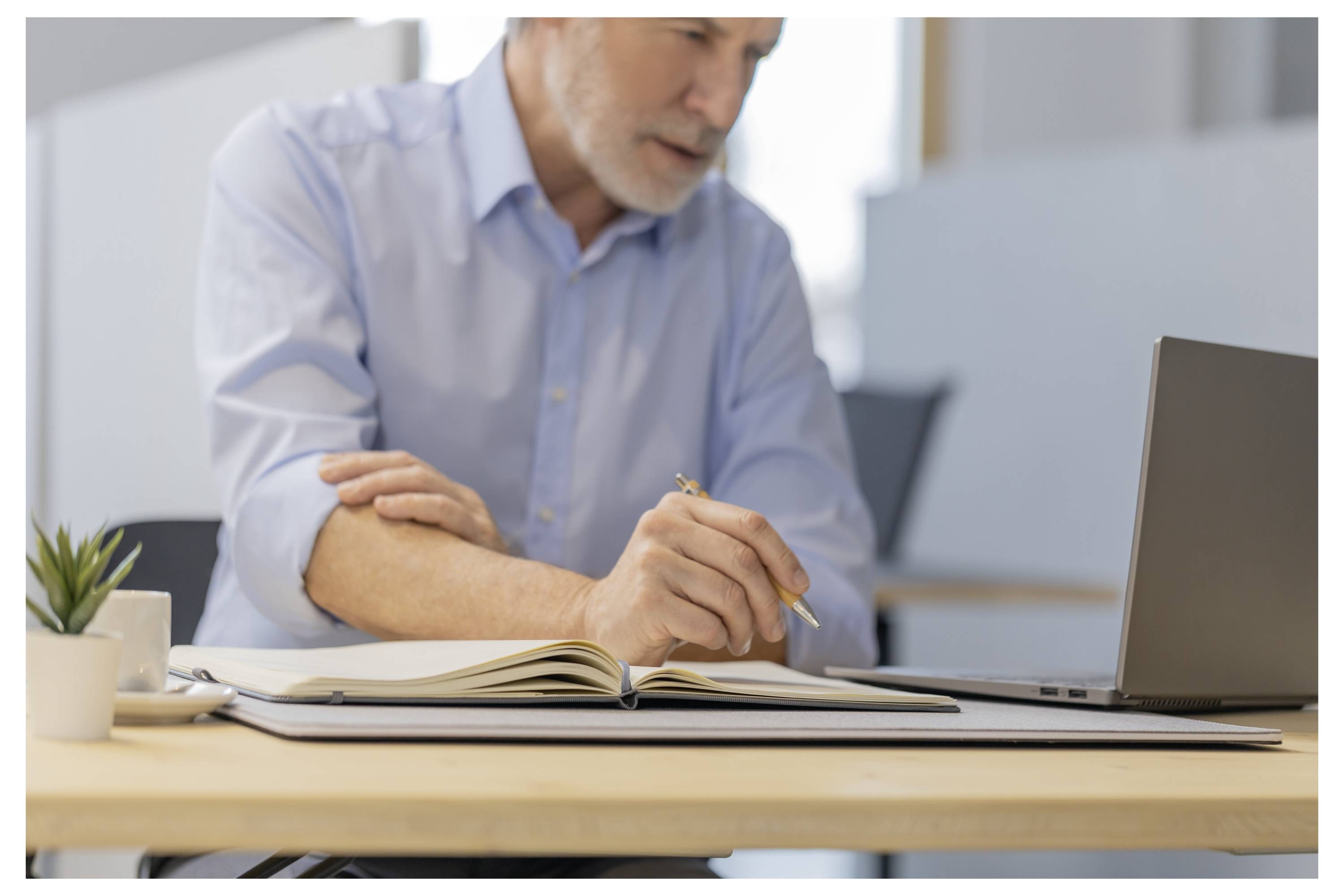 Uomo di mezza età con una camicia blu scrive in un quaderno accanto a un laptop su una scrivania, lavorando in un ufficio moderno.