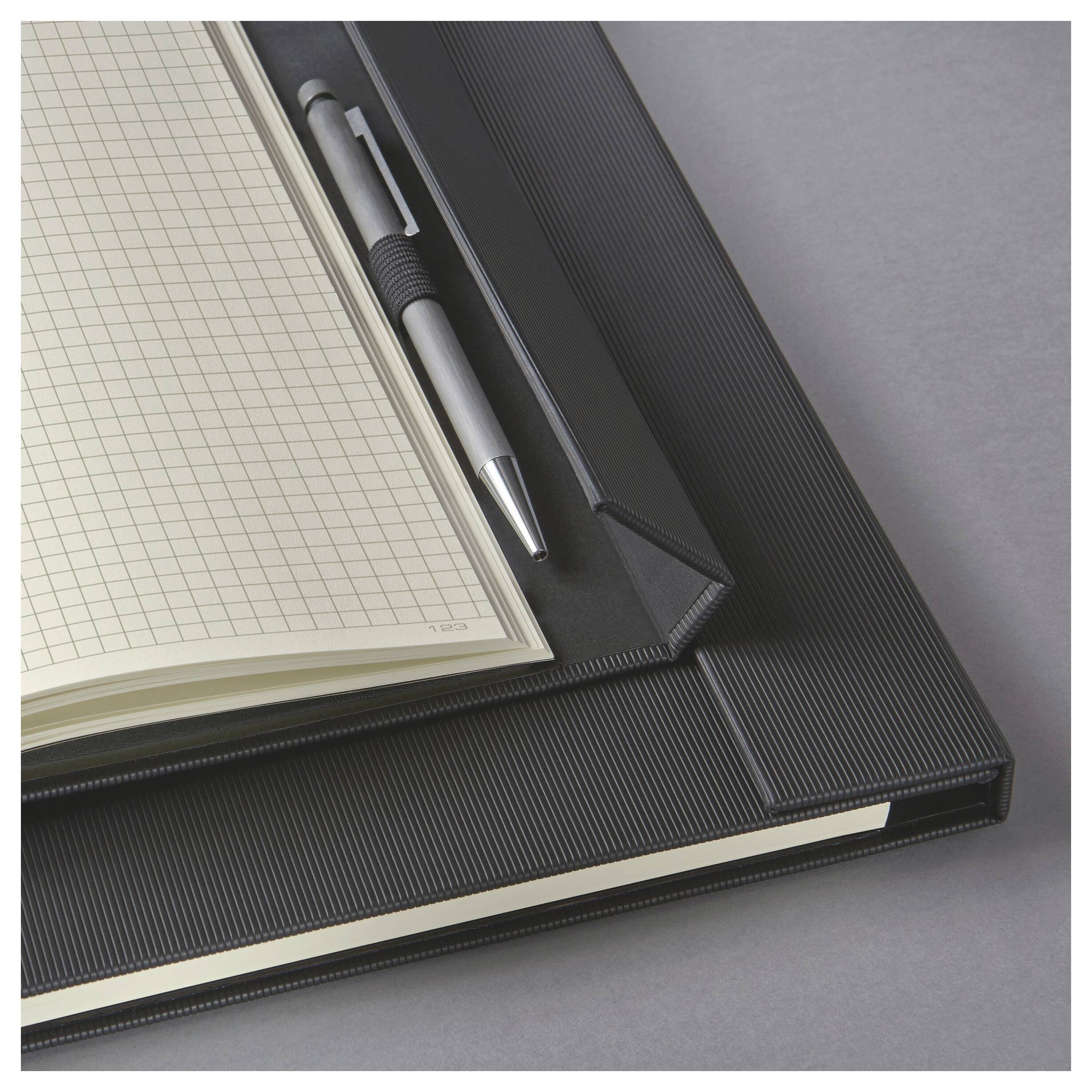 Quaderno aperto con pagine vuote a quadretti e una penna su una copertina nera con texture, mostrato su una superficie grigia.