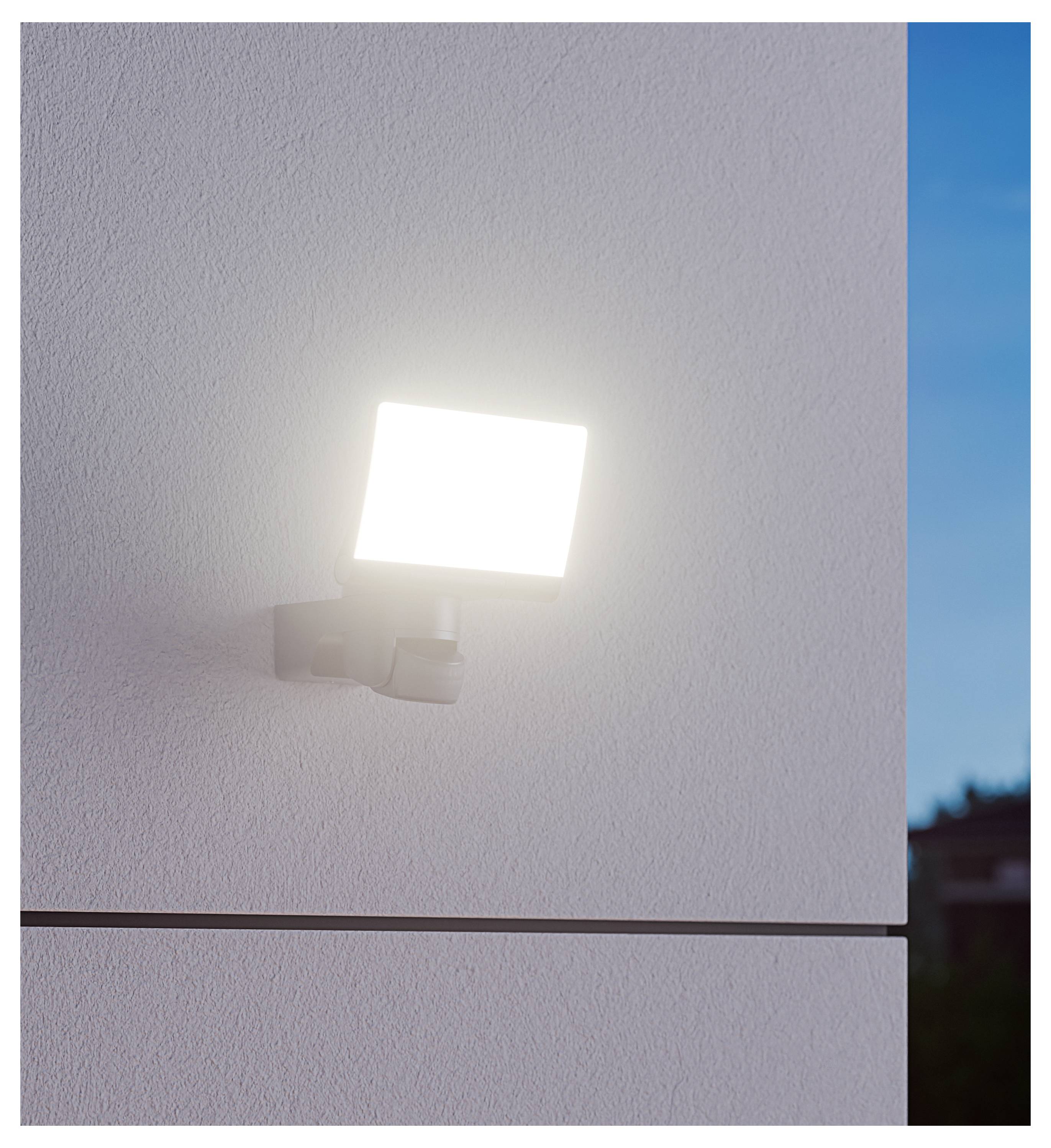 Apparecchio di illuminazione esterno montato a parete che illumina la facciata di un edificio bianco strutturato al crepuscolo.