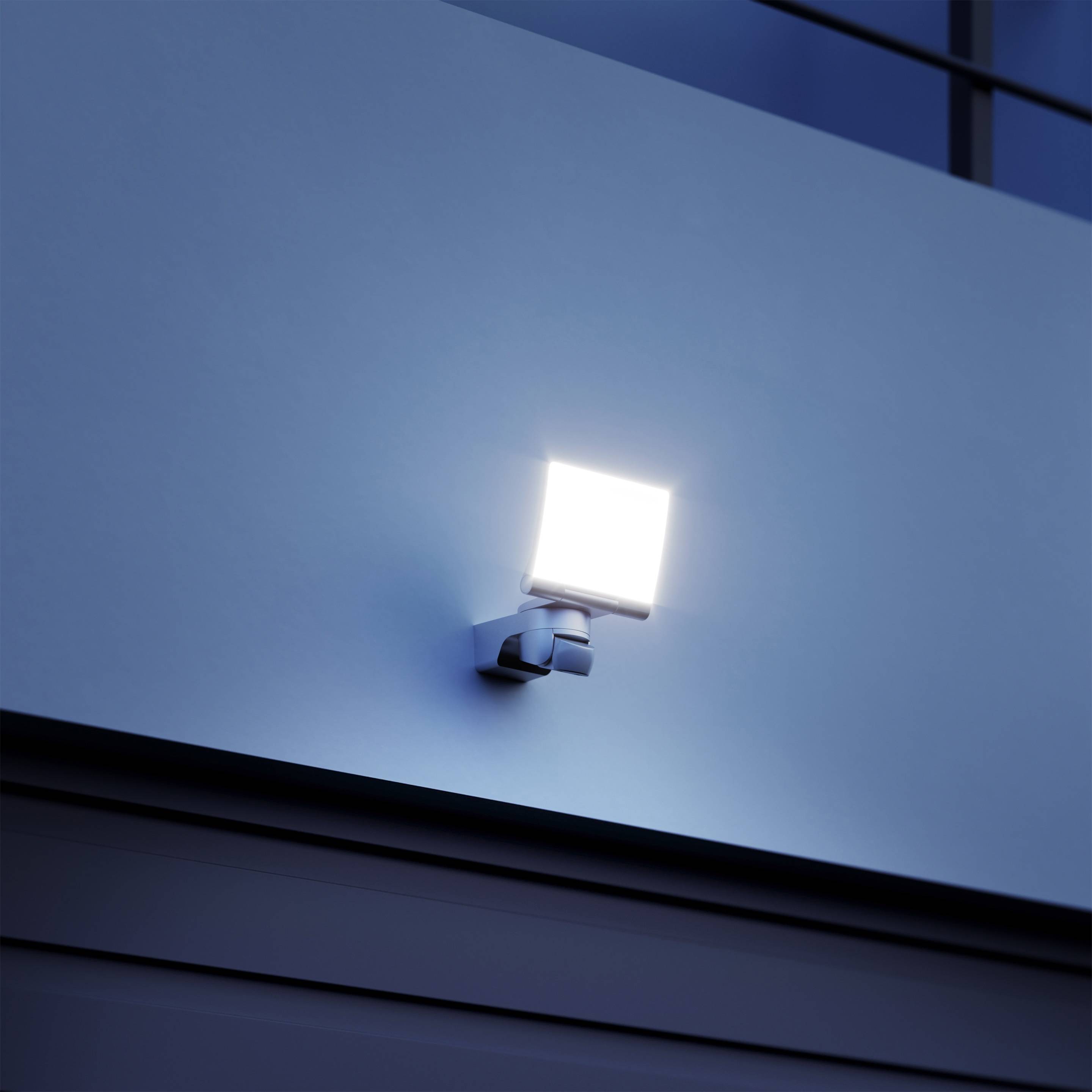 Una lampada rettangolare illumina una parete blu durante il crepuscolo. La lampada emana una luce bianca e brillante.
