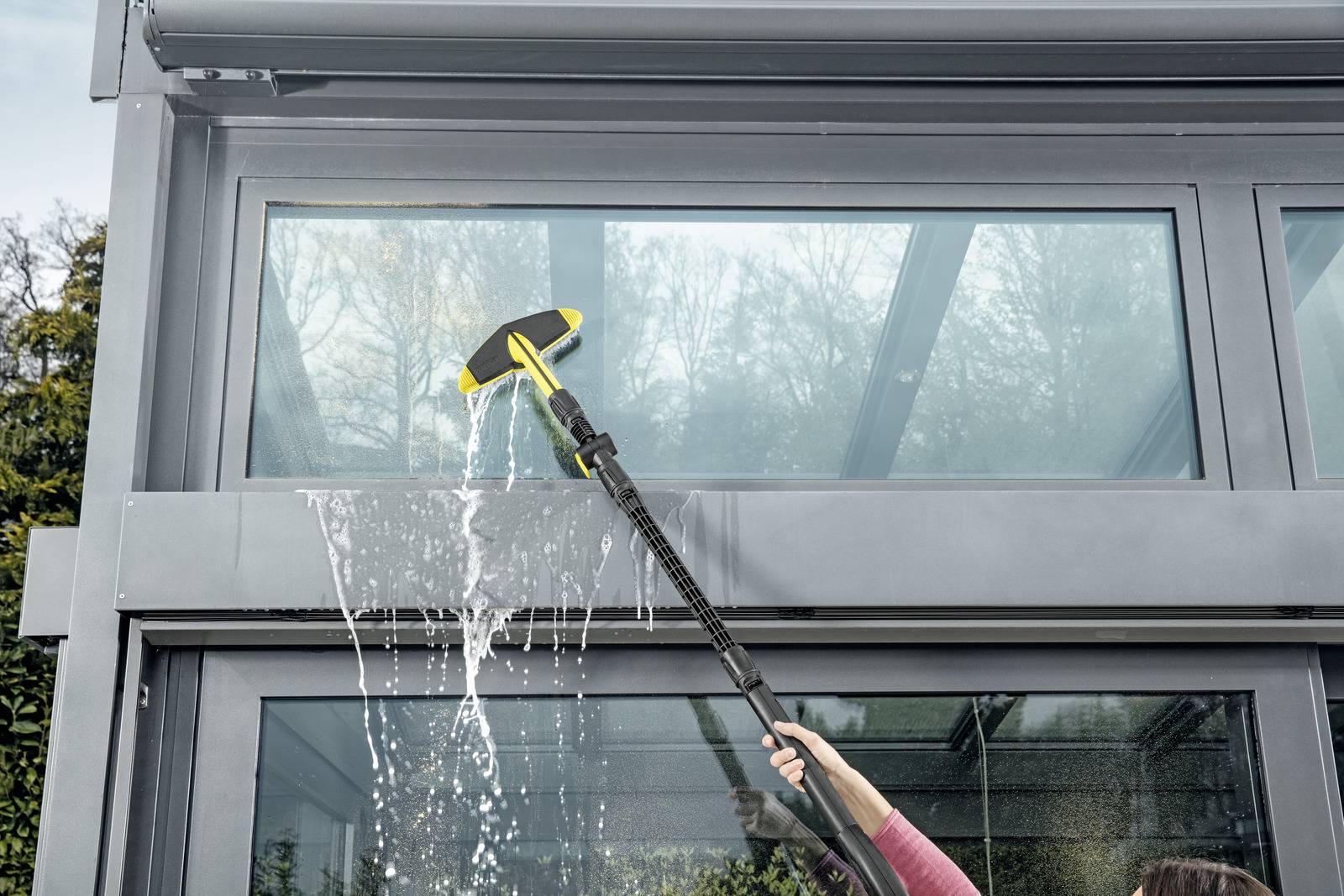 Una persona utilizza un attrezzo con manico lungo dotato di un accessorio per la pulizia per lavare un grande vetro di un edificio, con spruzzi d'acqua visibilmente schizzanti.