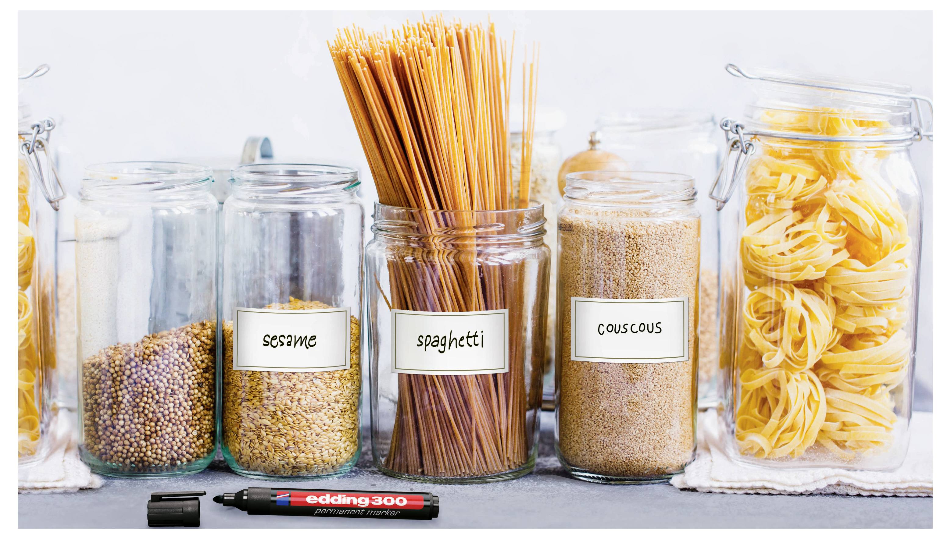 Cinque barattoli di vetro con alimenti secchi su uno scaffale, etichettati sesamo, spaghetti, cuscus e pasta, con un pennarello senza tappo davanti.