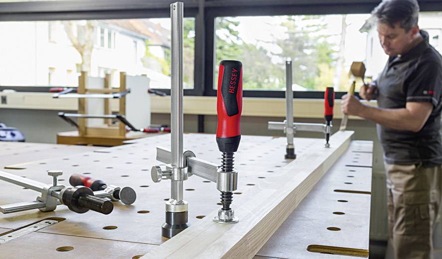 Un uomo lavora a un banco da lavoro in legno con diverse morse. Sullo sfondo ci sono grandi finestre che danno verso l'esterno.