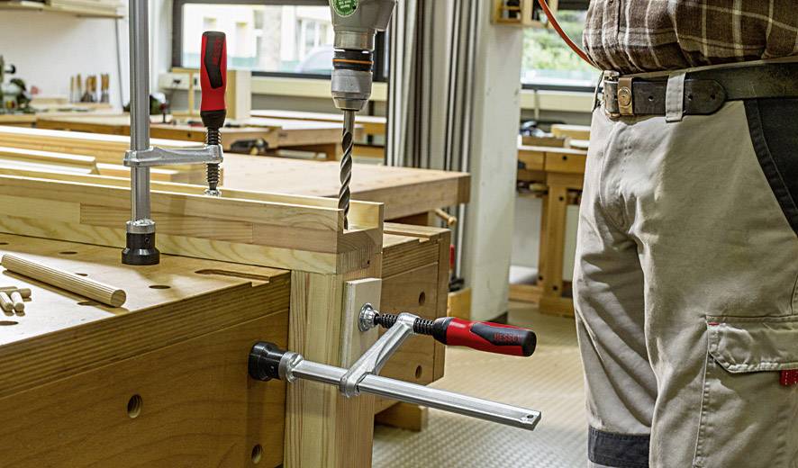 Una persona sta trapanando un pezzo di legno con un trapano, fissato a una scrivania in un laboratorio con delle morse.