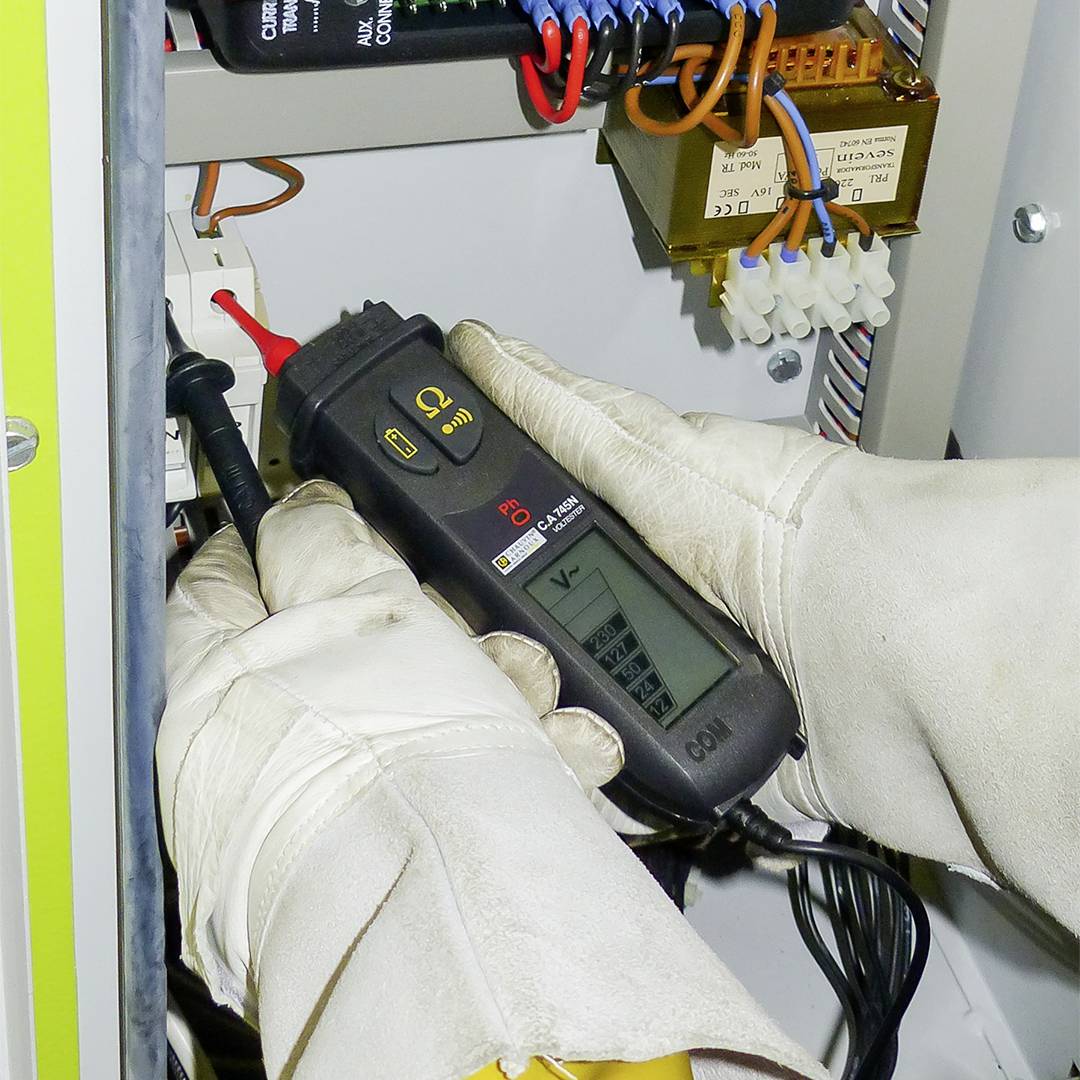 Un tecnico misura la tensione elettrica in un quadro elettrico utilizzando un rilevatore di tensione. Cavi e componenti sono visibili.
