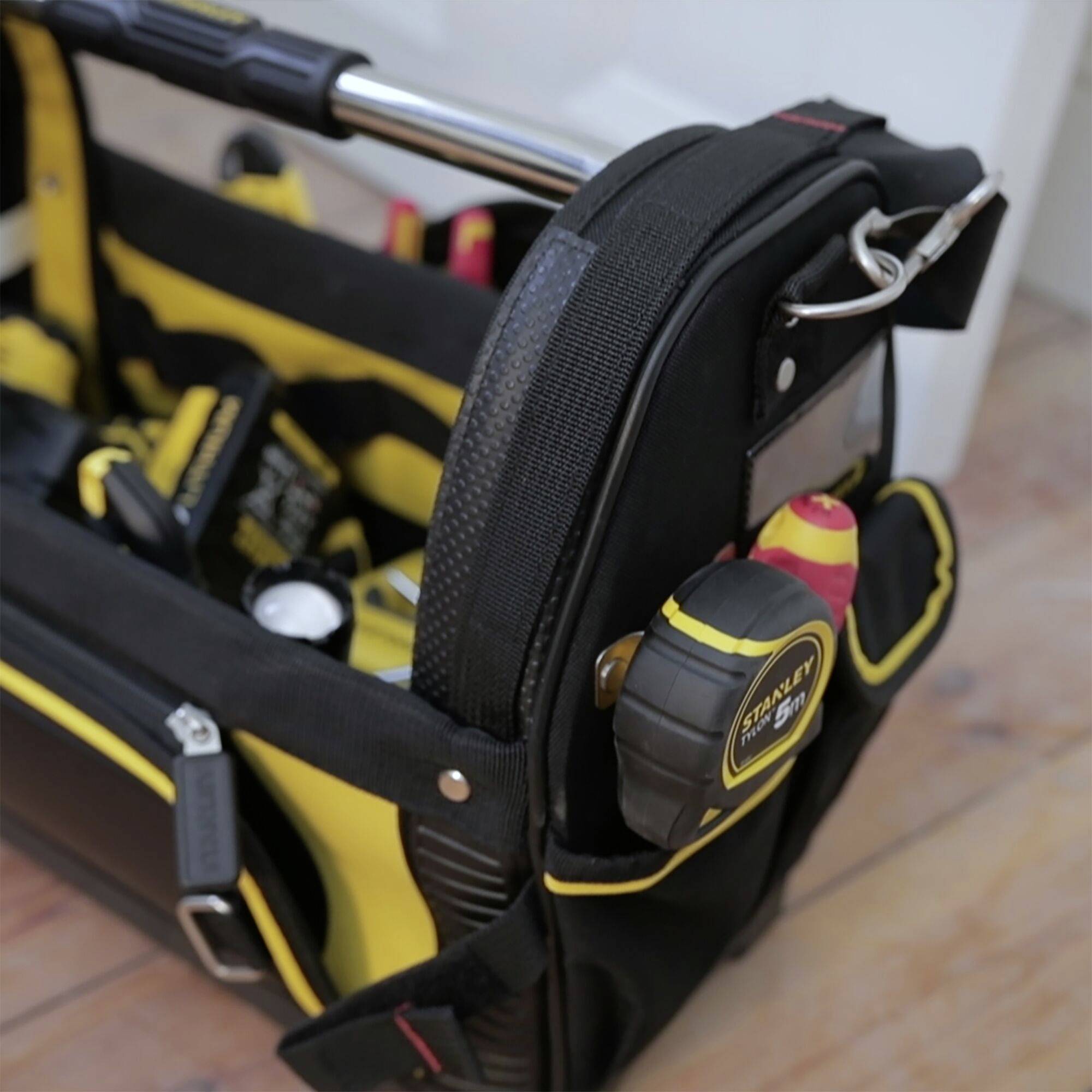 Borsa degli attrezzi con design giallo e nero su un pavimento di legno. Contiene diversi utensili manuali come nastro metrico, cacciavite e pinze.