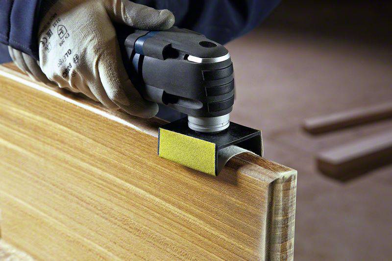 Persona che sta levigando i bordi di una tavola di legno con un utensile oscillante. La tavola è posizionata su un banco da lavoro in un laboratorio.