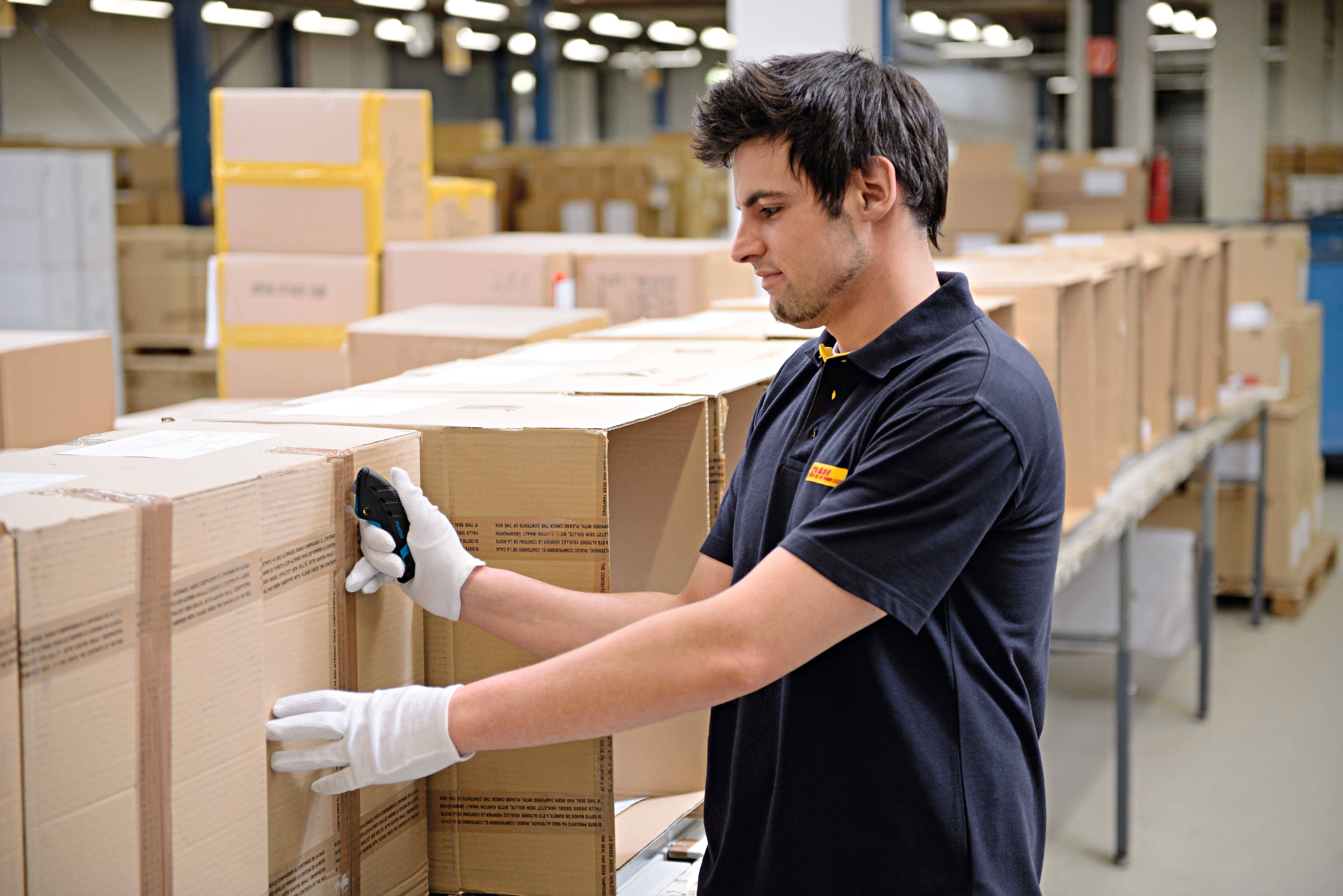 Un uomo scansiona pacchi in un magazzino. Indossa guanti e abbigliamento da lavoro. Sullo sfondo sono visibili numerosi scatoloni.