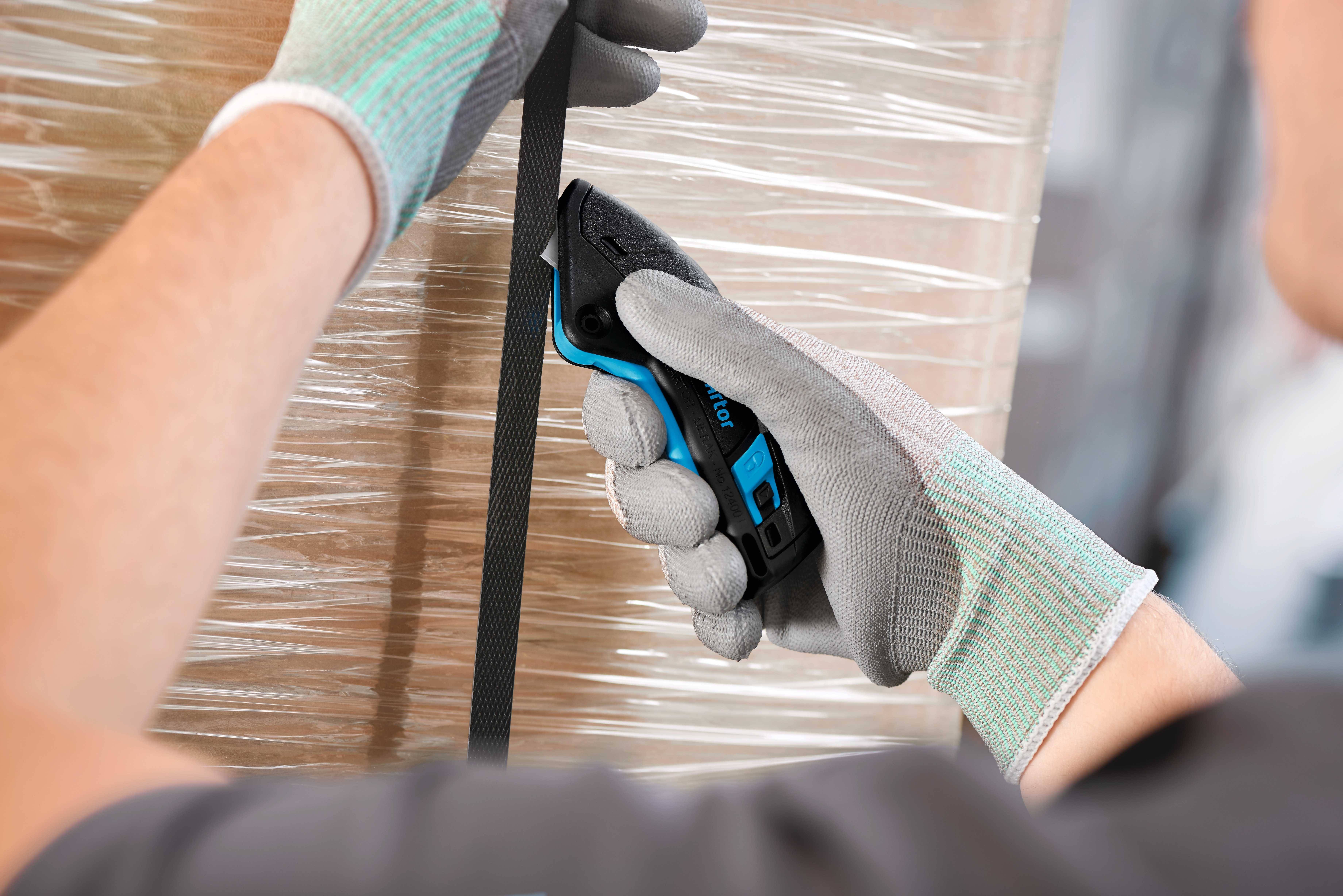 Una persona con guanti grigi usa un taglierino di sicurezza per tagliare una fascetta nera di plastica intorno a un imballaggio.