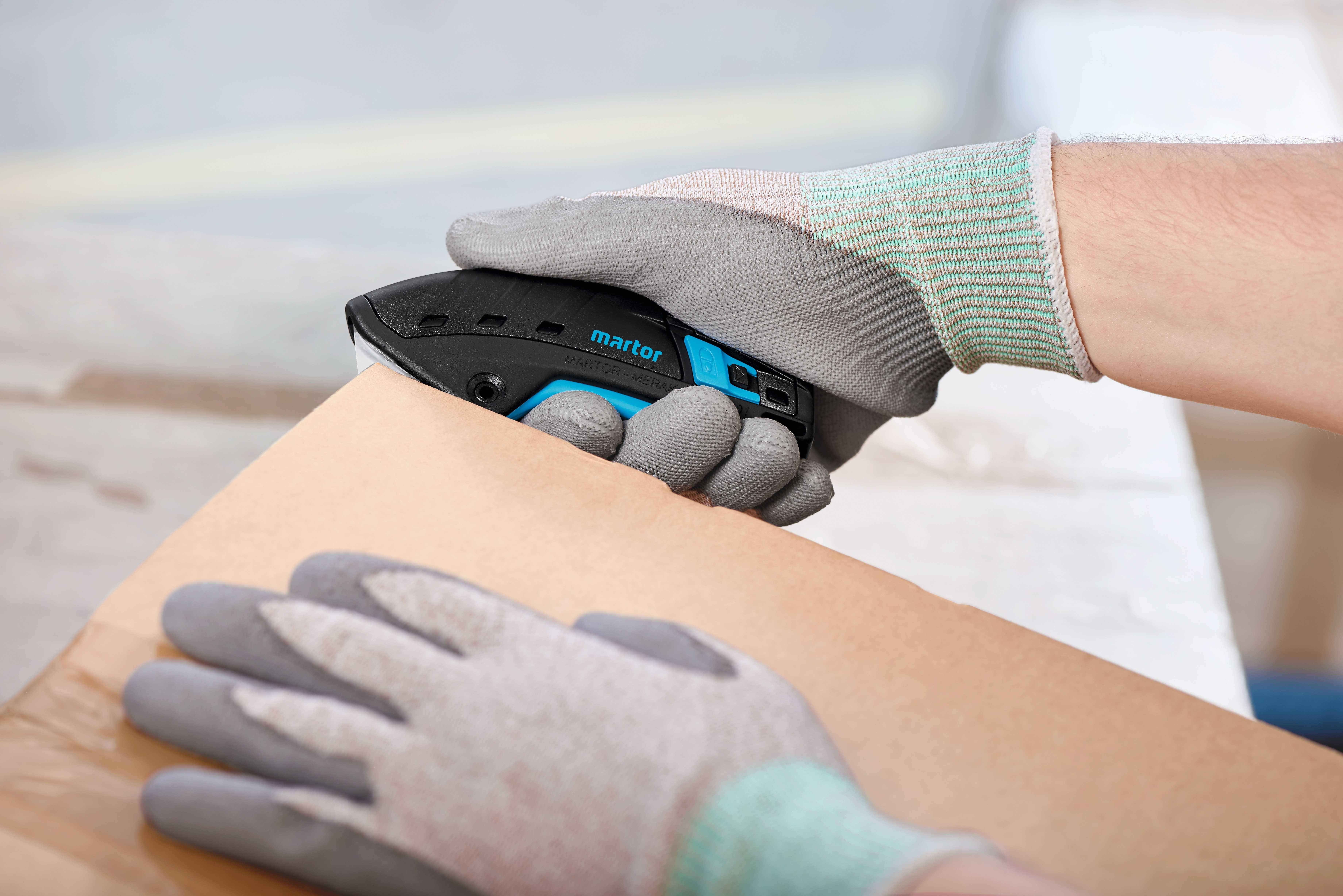 Persona con guanti protettivi taglia un pacco con un cutter su un banco da lavoro.