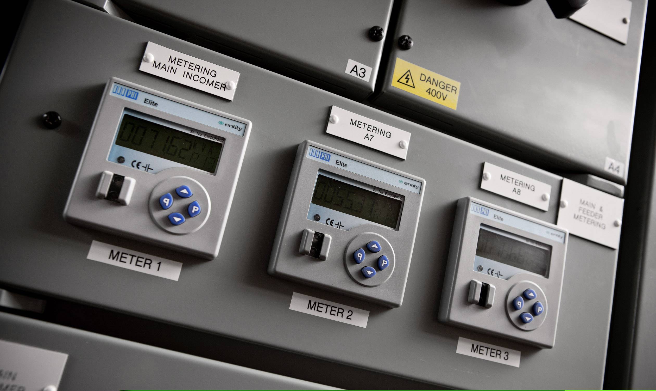 Tre contatori digitali dell'elettricità su un pannello di controllo grigio, sotto il testo 'Metering Main Incomer' e l'avviso di pericolo 'Danger 400V'.