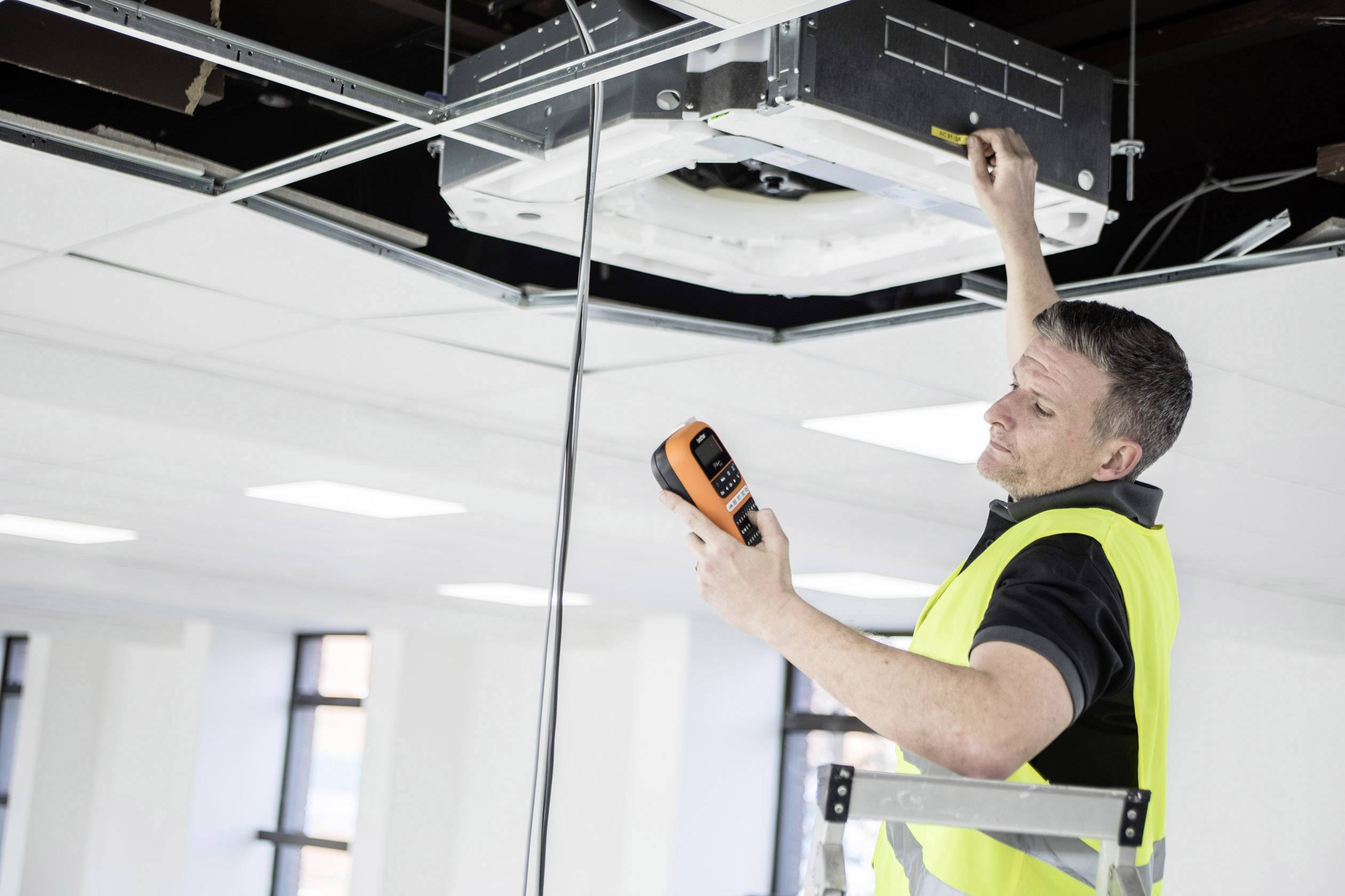 Un uomo con un giubbotto ad alta visibilità è in piedi su una scala, controlla un impianto di condizionamento sul soffitto con uno strumento di misurazione. Tiene in mano un attrezzo.
