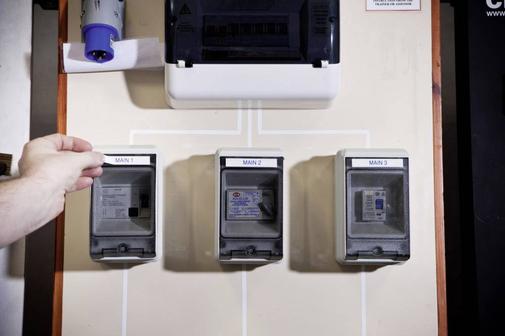 Tre interruttori elettrici con le etichette 'MAIN 1', 'MAIN 2' e 'MAIN 3'. Una mano aziona l'interruttore 'MAIN 1'.