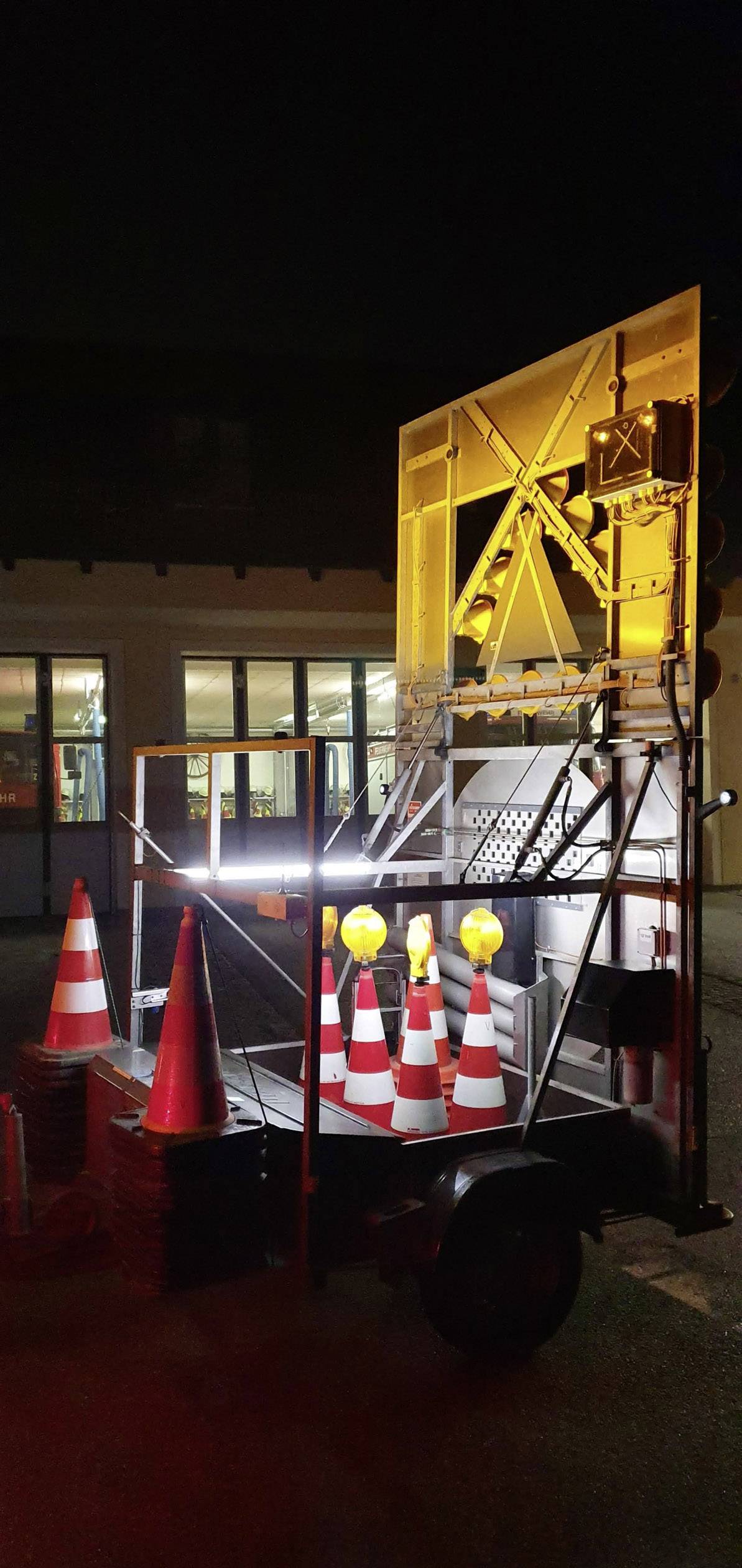 Un rimorchio luminoso per la sicurezza stradale con diversi coni segnaletici e luci lampeggianti gialle durante la notte, in un contesto urbano.