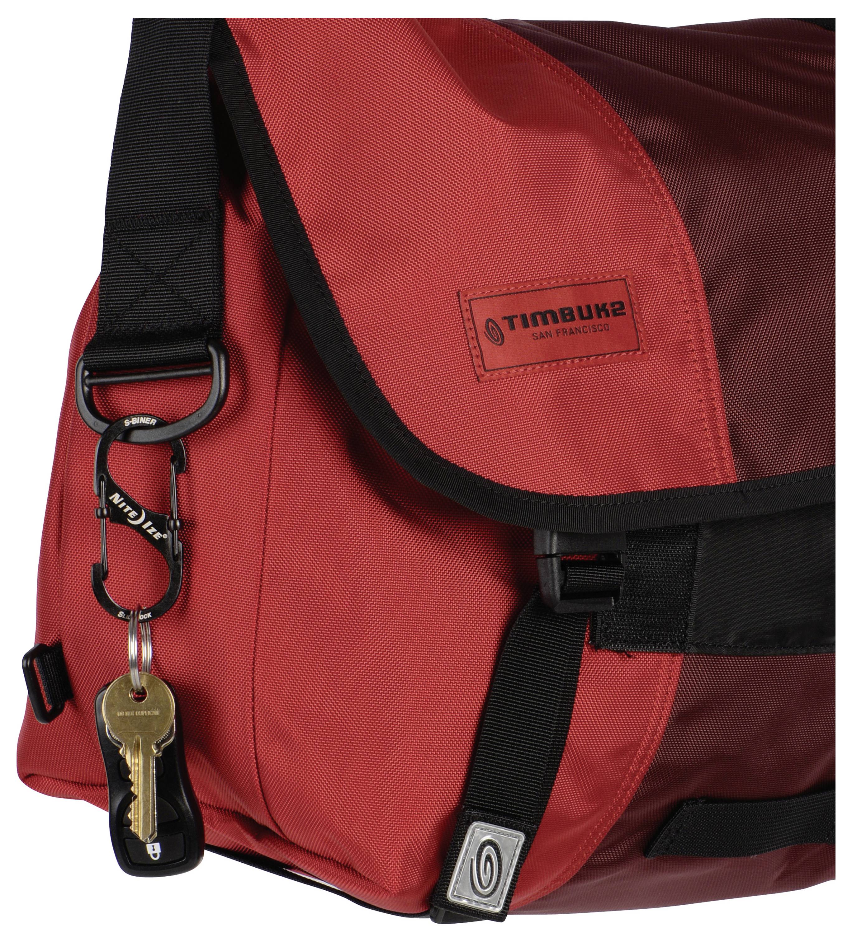 Una borsa messenger rossa con una tracolla nera, con il logo 'Timbuk2 San Francisco', e un portachiavi appeso a una fibbia anteriore.