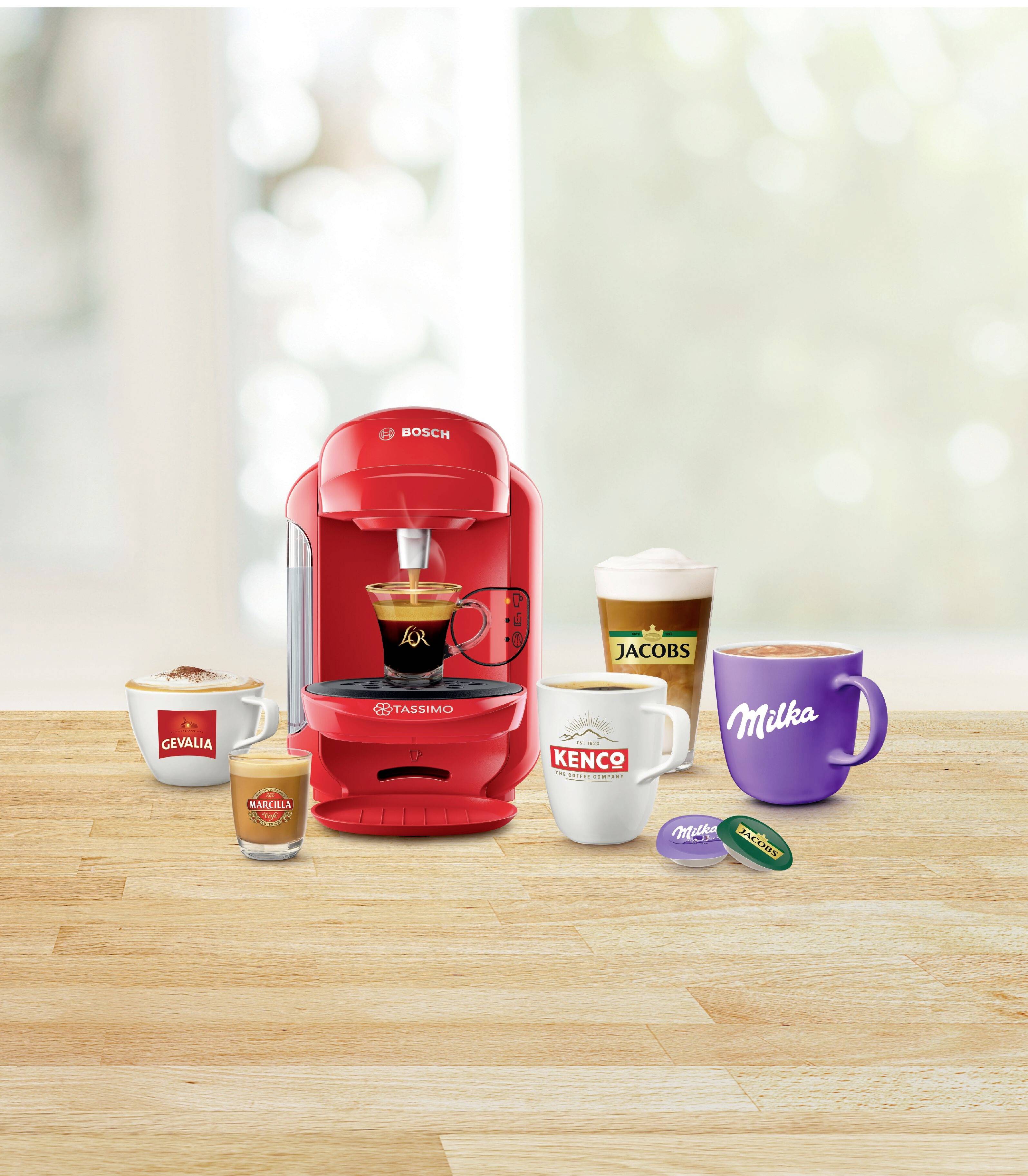 Macchina rossa per capsule di caffè con quattro tazze a marchio 'Jacobs', 'Milka', 'Kenco' e 'Caffè Onyx'. Capsule di caffè disposte accanto sulla tavola di legno.