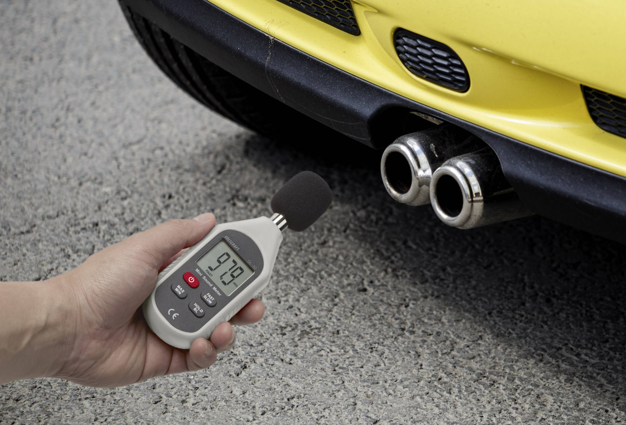 Una mano tiene un misuratore di decibel vicino a un tubo di scarico giallo e rumoroso di un'auto. Lo strumento indica 97,9 decibel.