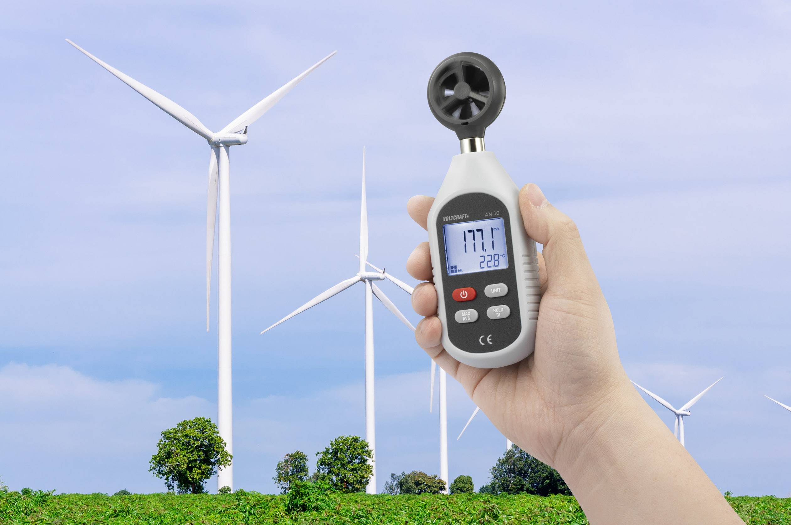Una mano tiene un anemometro con display digitale, mostrato davanti a turbine eoliche in un campo verde sotto un cielo blu.