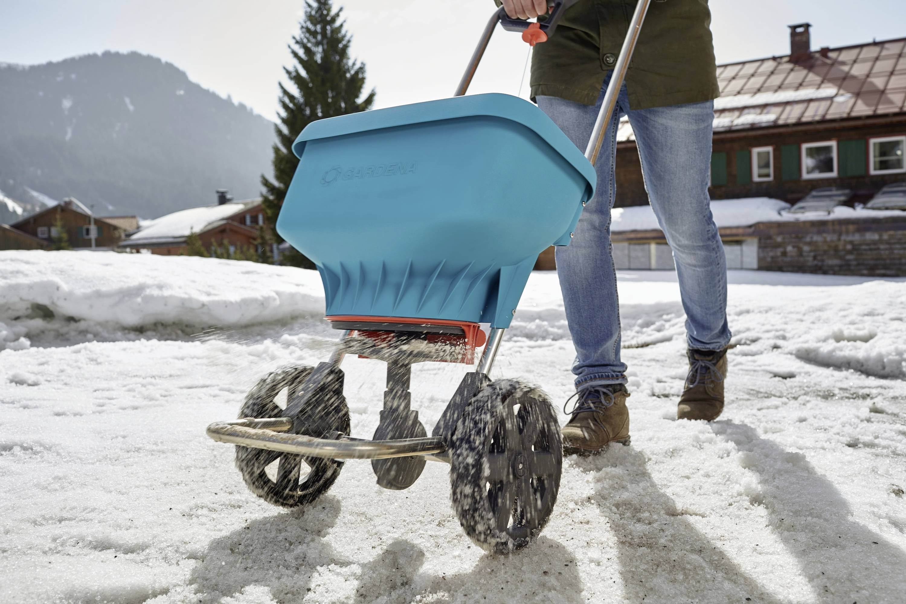 Una persona sparge sale su un terreno innevato con un carrello spargi-sale blu. Sullo sfondo si vedono case e una montagna con tempo soleggiato.