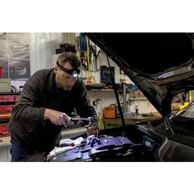Un meccanico lavora con una torcia elettrica sul motore aperto di un'auto in un'officina. Attrezzi e attrezzature sono visibili sullo sfondo.