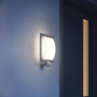 Una moderna applique è illuminata accanto a una porta. La lampada ha un design quadrato e irradia una luce delicata e calda.