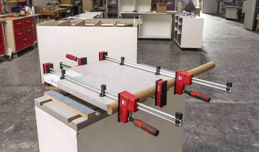 Una tavola di legno viene fissata su un banco da lavoro con grandi morsetti rossi. Sullo sfondo si intravede l'ambiente di un laboratorio con armadi e attrezzi.