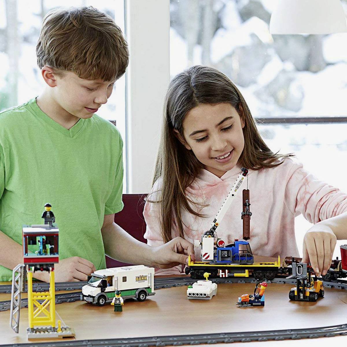 Due bambini giocano con un set di treni Lego su un tavolo, composto da un treno merci, binari e edifici.