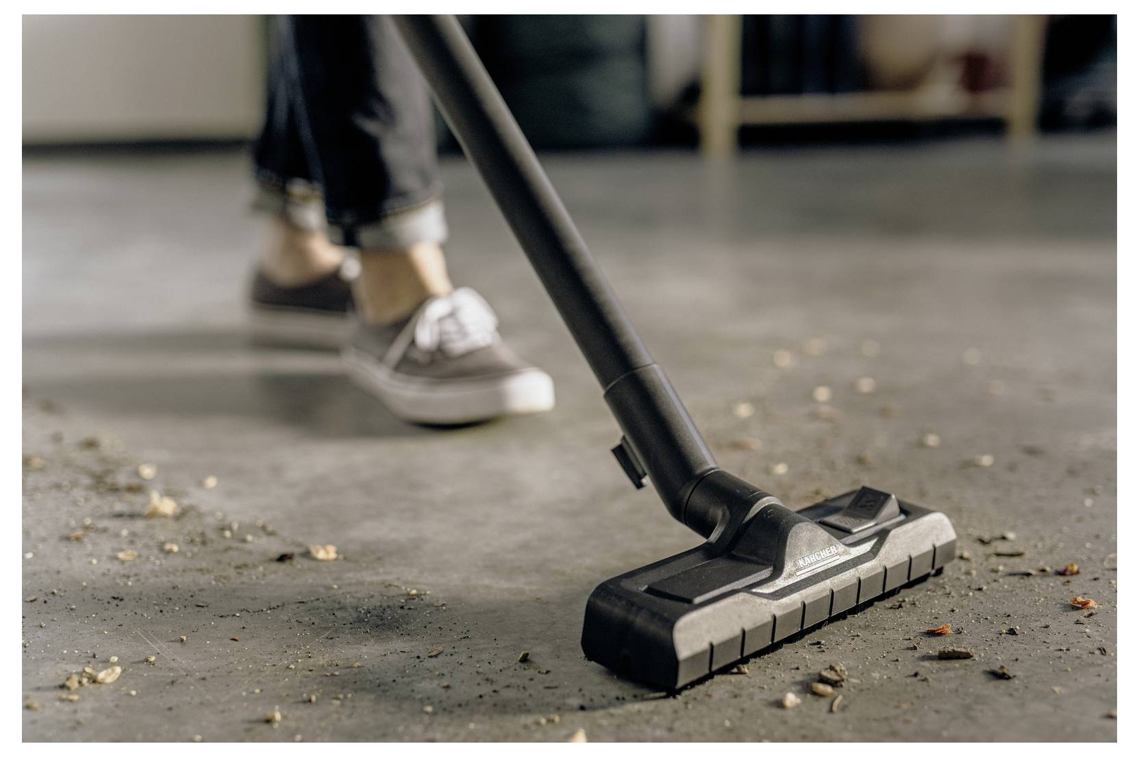 Una persona che aspira un pavimento cosparso di detriti, concentrandosi sulla testina dell'aspirapolvere mentre pulisce, indossando scarpe casual e jeans.