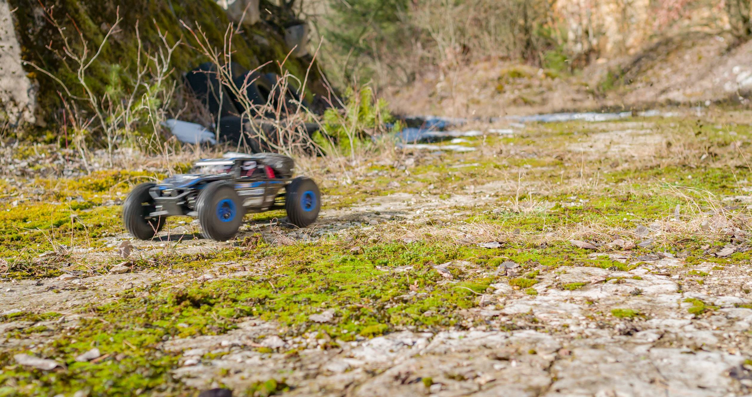 Un'auto telecomanda sfreccia velocemente su un sentiero muschiato e sassoso in un'area boschiva. Alberi e cespugli sullo sfondo.
