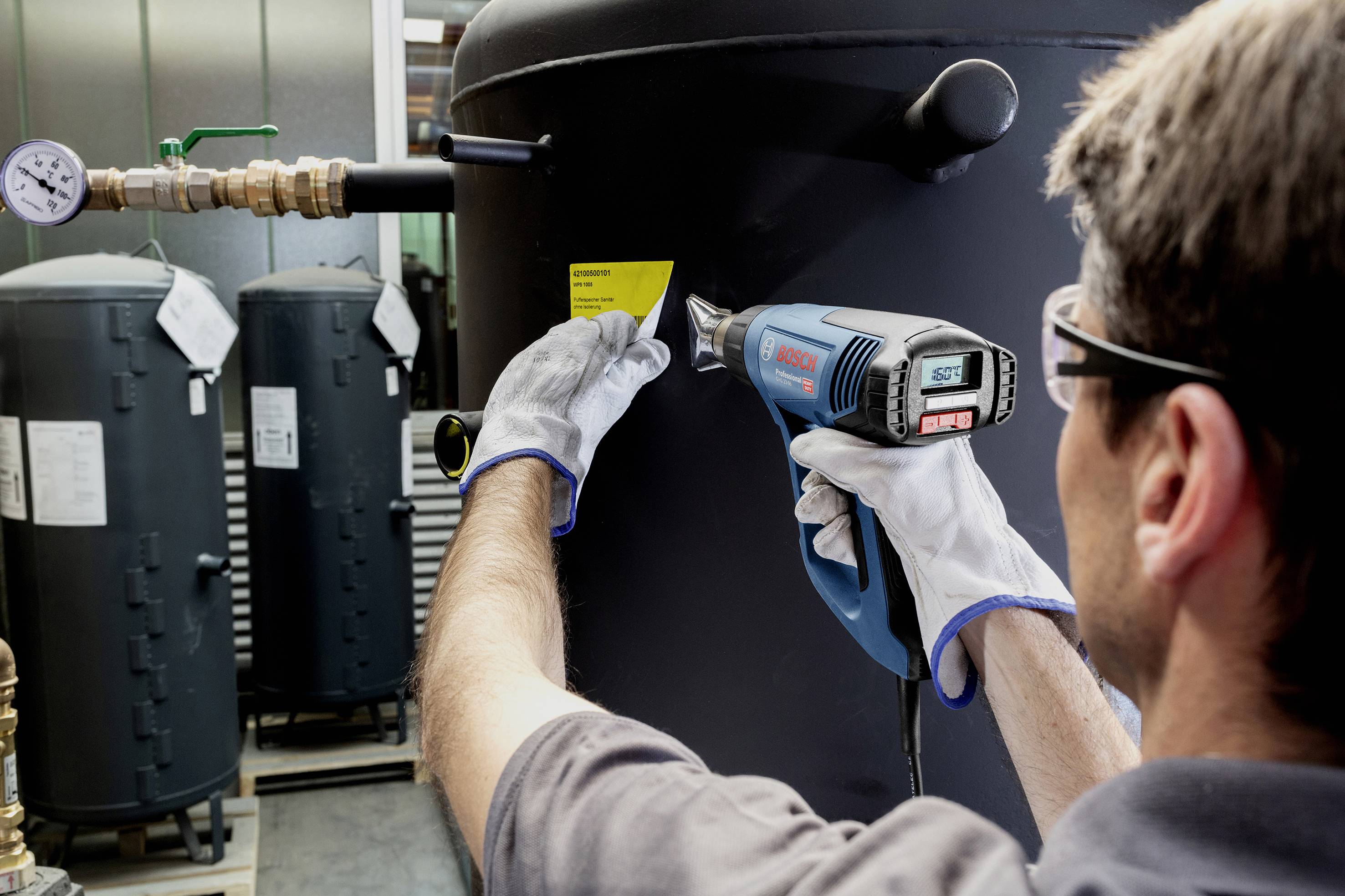 Una persona con abbigliamento da lavoro sta utilizzando un soffiatore ad aria calda per applicare un'etichetta gialla su un grande contenitore nero.