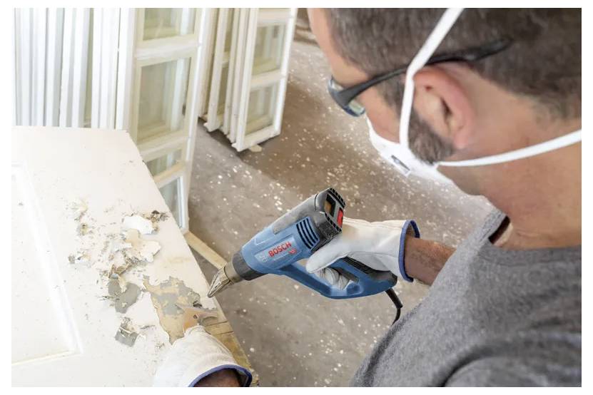 Una persona che indossa guanti e una mascherina usa un cannello termico per rimuovere la vernice da una superficie in legno, con finestre impilate sullo sfondo.