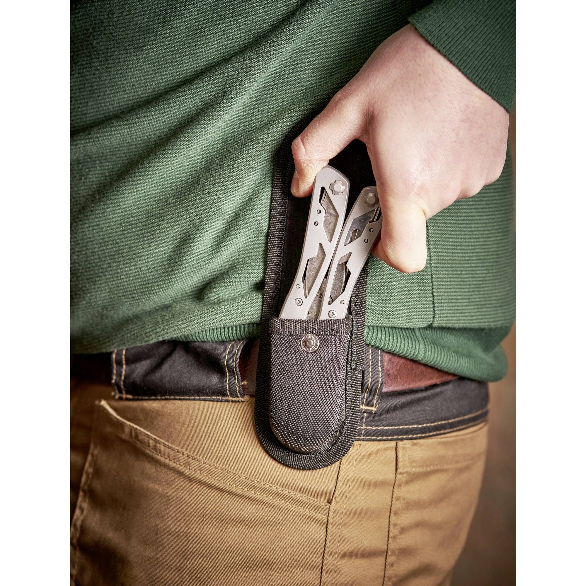 Una persona infila un multiuso in un fodero sulla cintura sul lato destro del corpo. Indossa una maglia verde e pantaloni marroni.