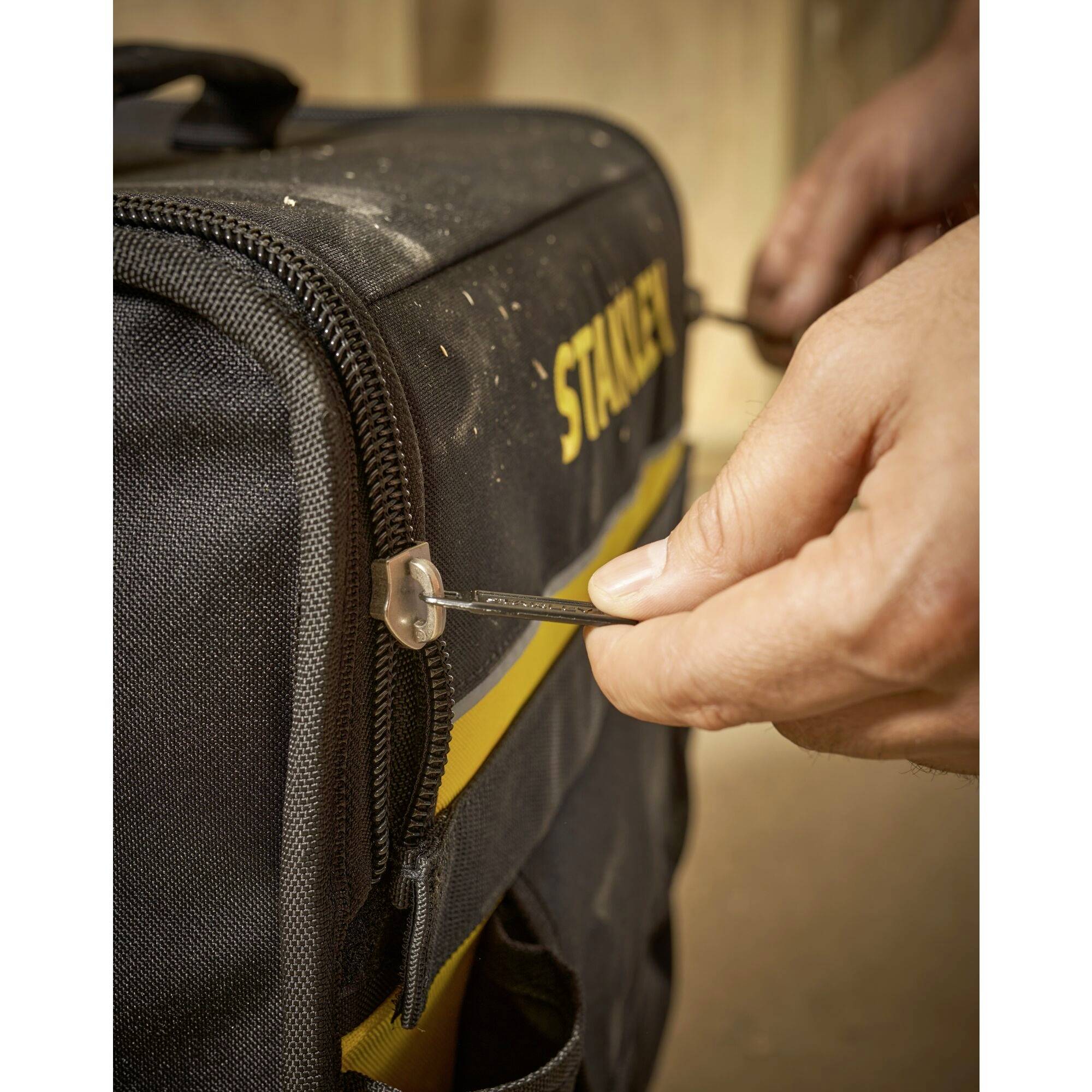 Una mano tira la zip di una borsa degli attrezzi nera. La scritta gialla recita 'STANLEY'.