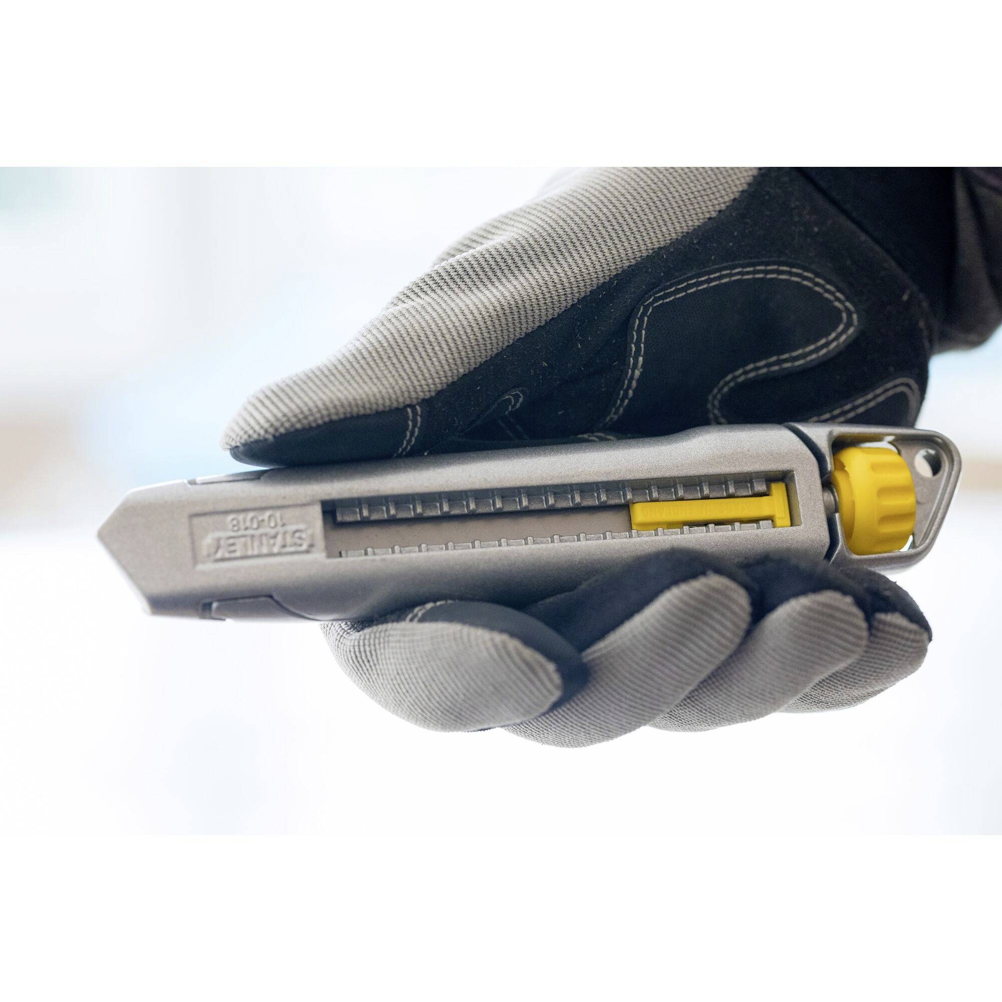 Una mano tiene un taglierino per tappeti con lama giallo-grigia e scala graduata in un ambiente luminoso. Il coltello è parzialmente aperto.