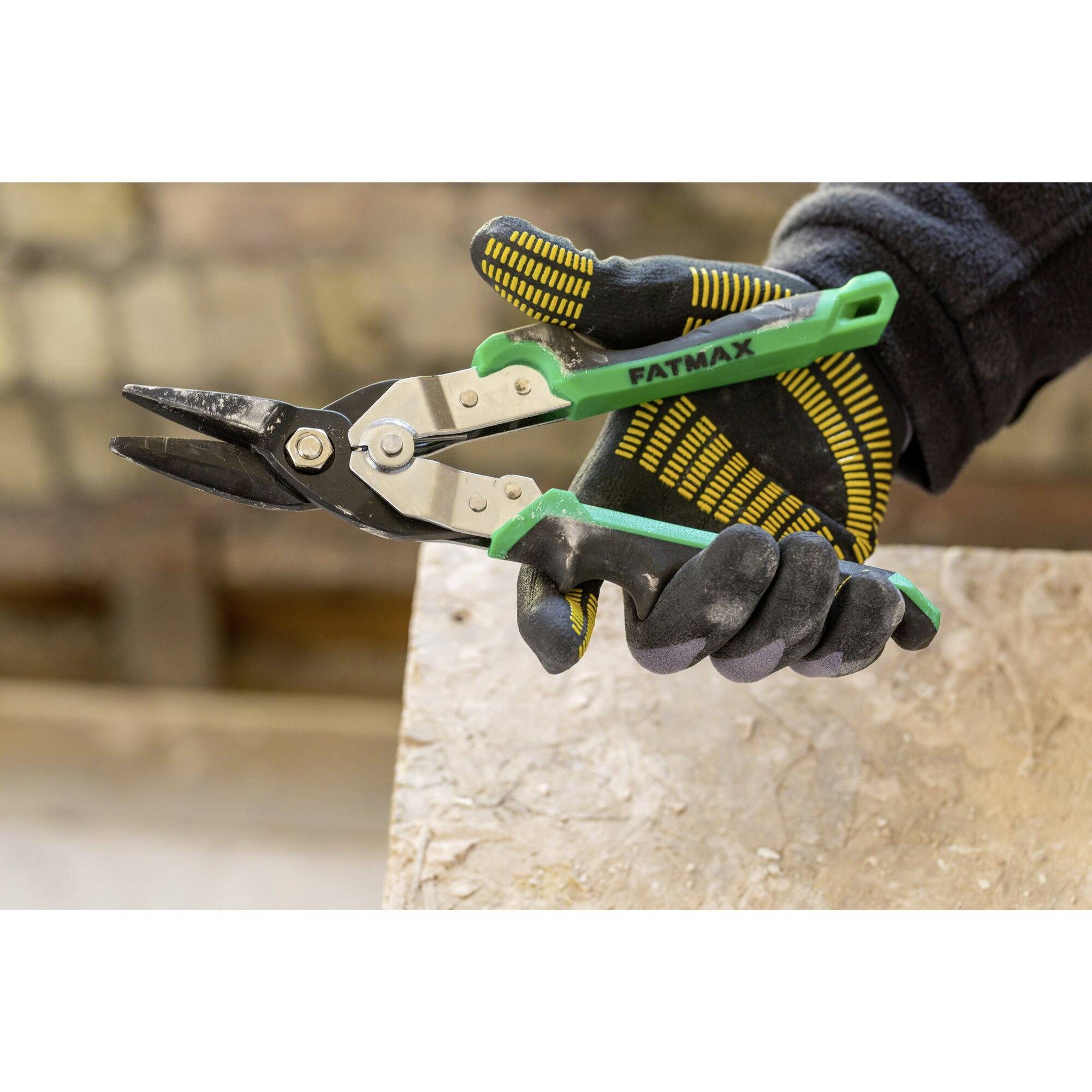 Una mano con un guanto da lavoro tiene una forbice verde per lamiera con la scritta 'FATMAX', sullo sfondo sfocato.