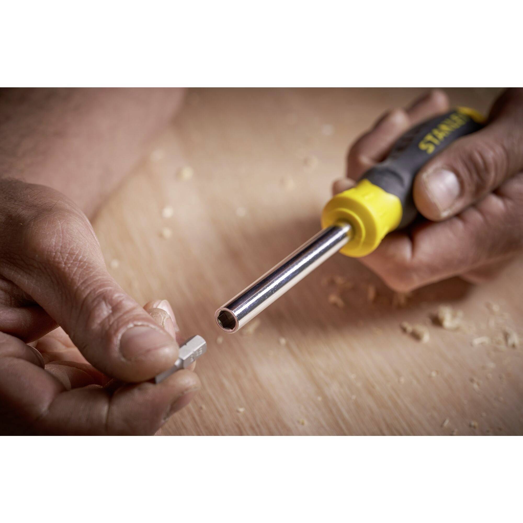 Una persona tiene un manico giallo e nero nella mano destra e un piccolo pezzo di metallo nella mano sinistra, sopra una superficie di legno.