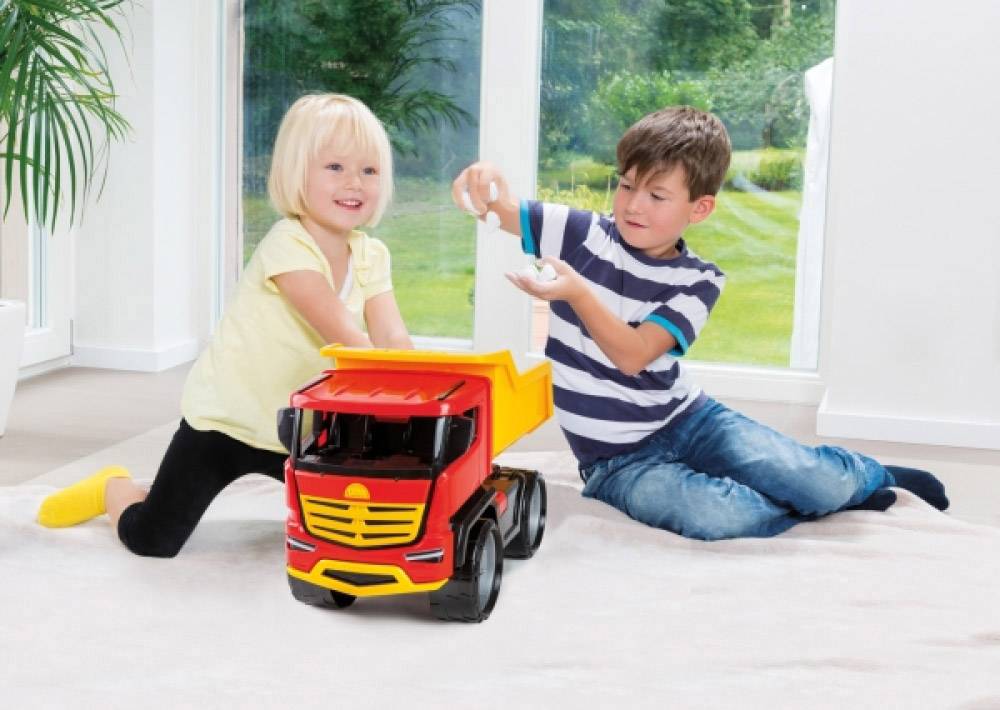 Due bambini giocano sul pavimento con un camion giocattolo rosso. Un bambino sta riempiendo qualcosa nel cassone, mentre la bambina osserva.