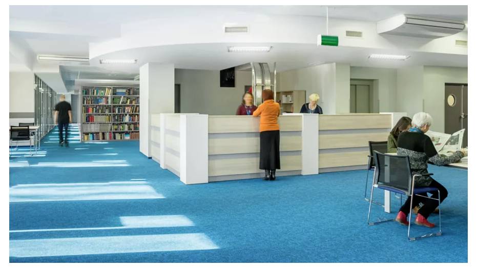 Un interno di biblioteca con un banco di prestito gestito da due persone. Una persona è in piedi al banco, mentre altri sfogliano e leggono libri.