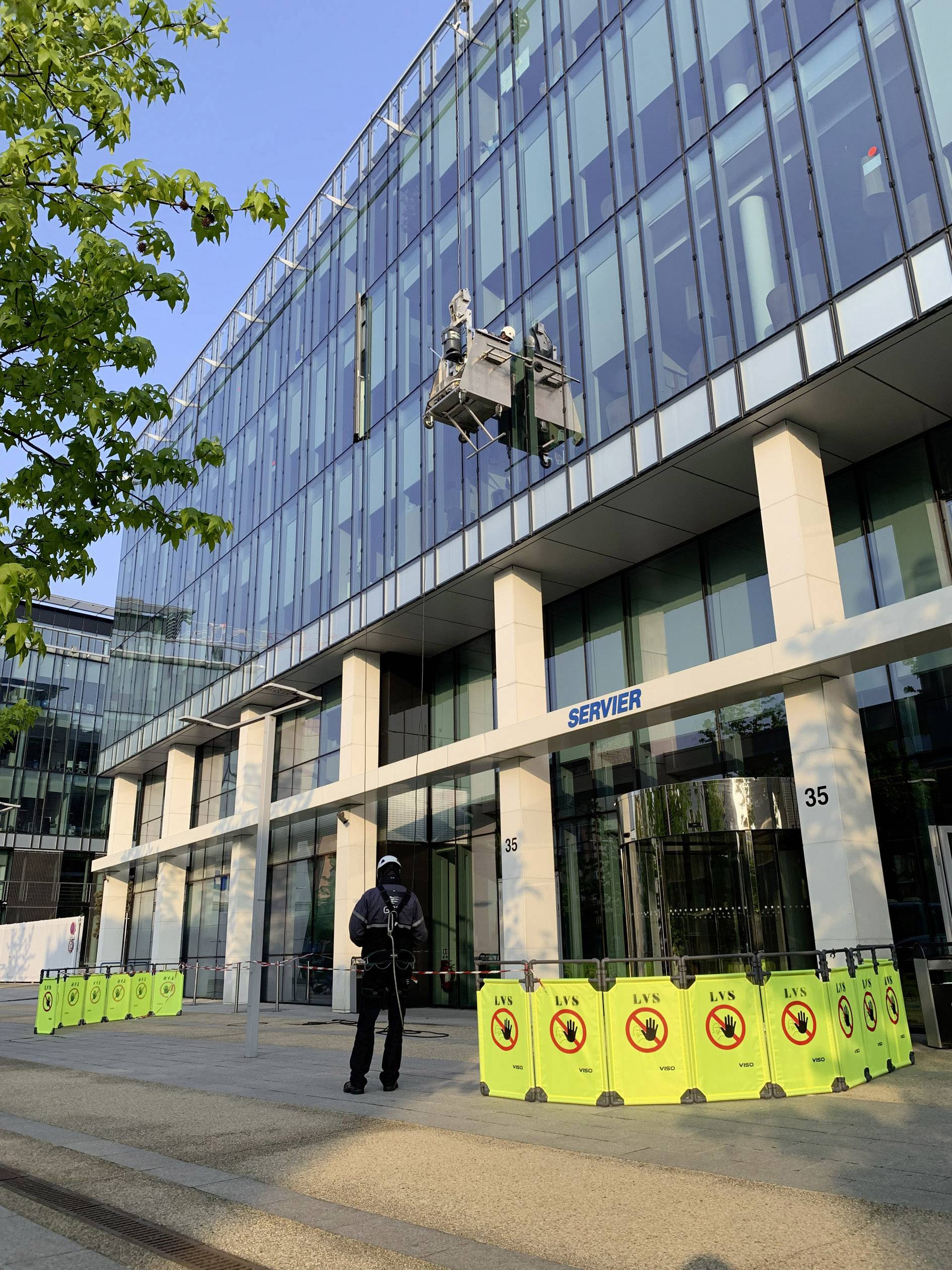 Un lavoratore su una piattaforma elevatrice pulisce la facciata vetrata di un moderno edificio per uffici. Nastri gialli di sicurezza delimitano l'area di lavoro sul fondo.