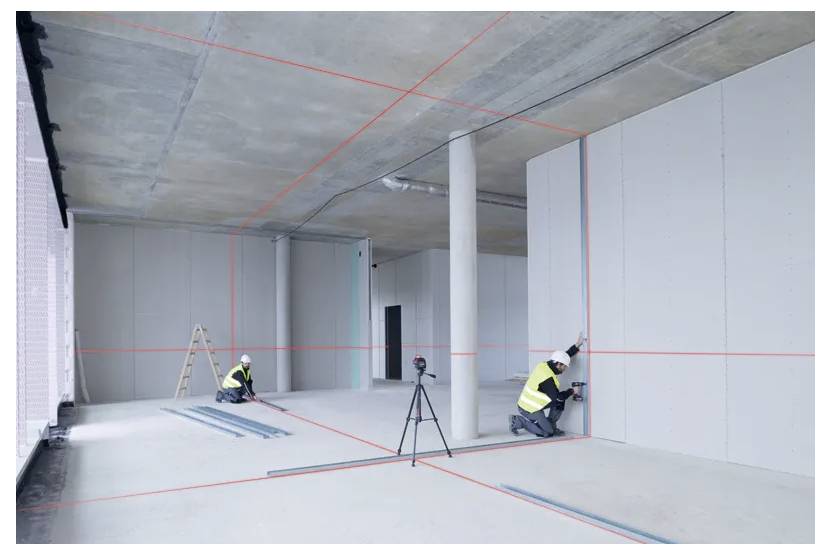 Due lavoratori edili con giubbotti di sicurezza utilizzano linee laser rosse per allineare pannelli di cartongesso in un interno di edificio non finito. Un treppiede è posizionato al centro.