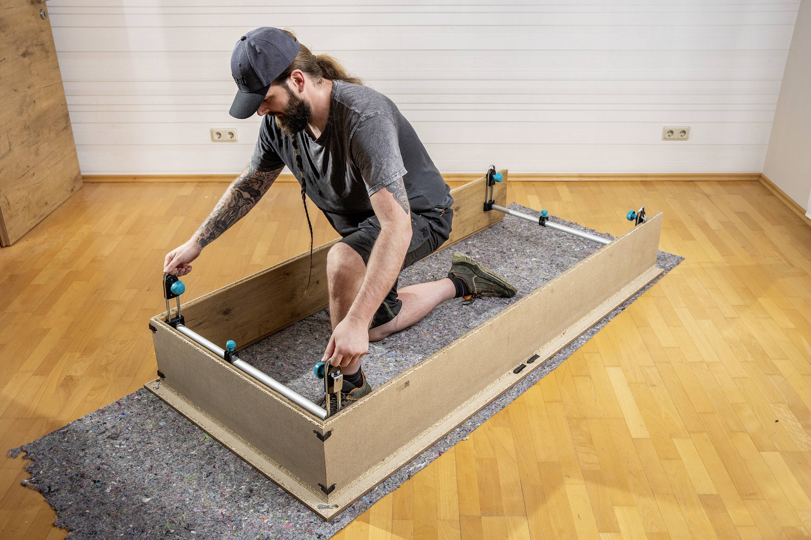 Un uomo inginocchiato, con degli attrezzi, sta assemblando un mobile in legno su un pavimento di legno. Sullo sfondo si trova una parete bianca.
