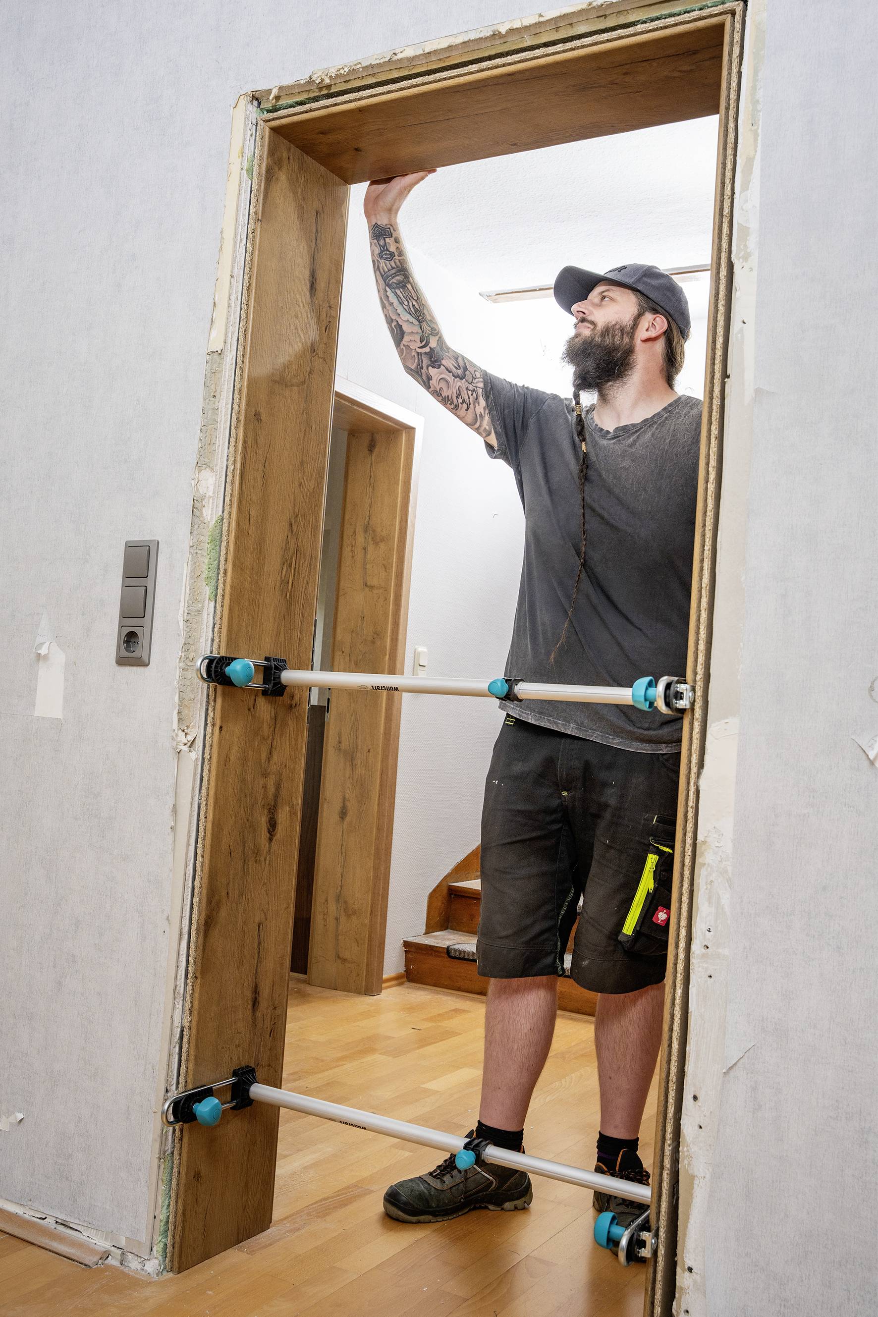 Un uomo sta montando un telaio porta in legno all'interno di una casa. Indossa abiti da lavoro e un cappello e utilizza attrezzature di supporto per l'installazione.