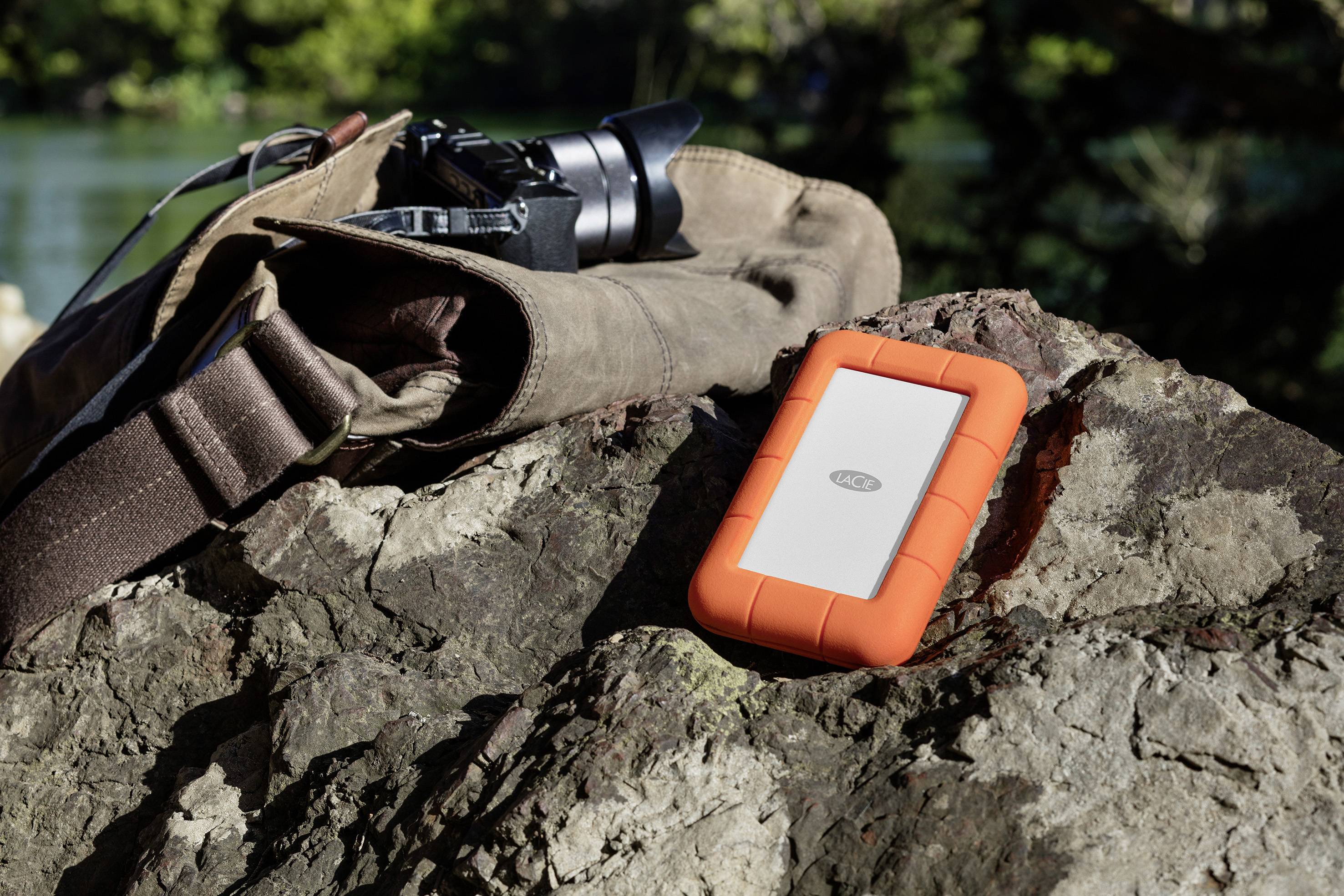 Una macchina fotografica e un hard disk arancione sono posizionati su una roccia. Sullo sfondo, sfocati, si vedono un bosco e un lago.