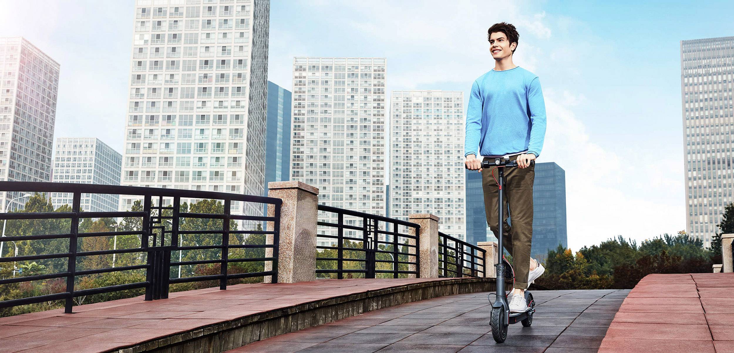 Una persona guida un monopattino elettrico su un ponte urbano, circondato da grattacieli moderni e un cielo blu.