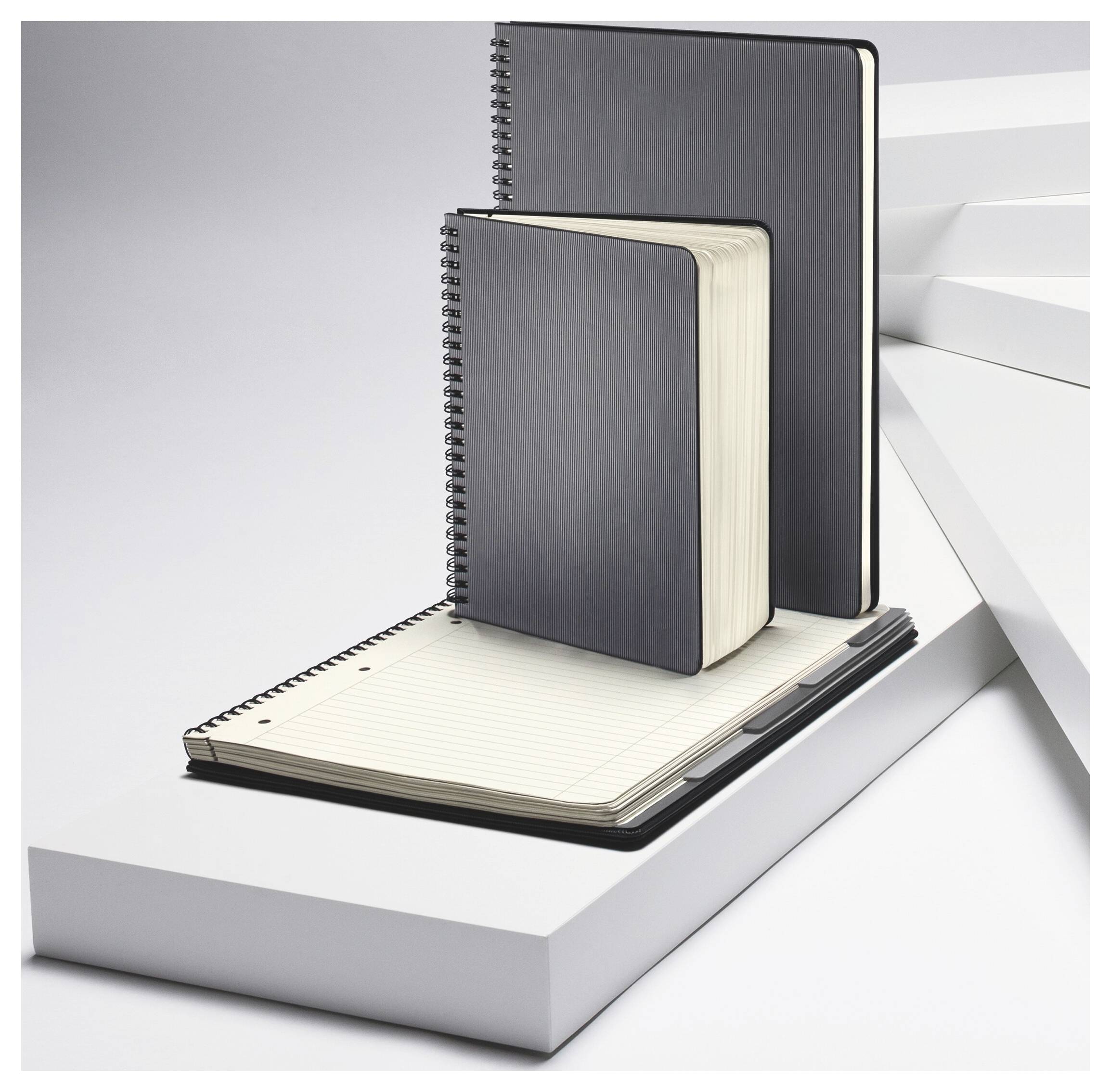 Tre quaderni rilegati a spirale impilati su un espositore bianco a gradini, con un quaderno aperto in bianco davanti.