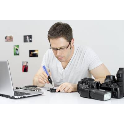 'Uomo ripara una fotocamera con un saldatore alla scrivania. Laptop e accessori per la fotocamera sul tavolo. Foto appese al muro sullo sfondo.'
