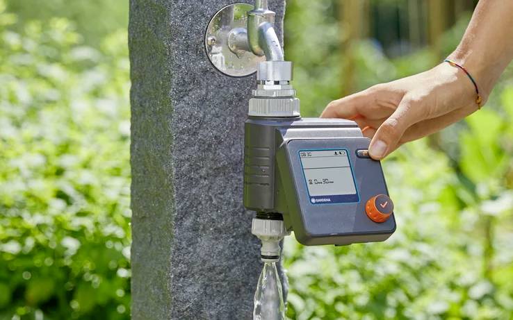 Una mano aziona un timer digitale per l'acqua collegato a un rubinetto, con uno sfondo di un giardino lussureggiante, che illustra l'irrigazione automatizzata.
