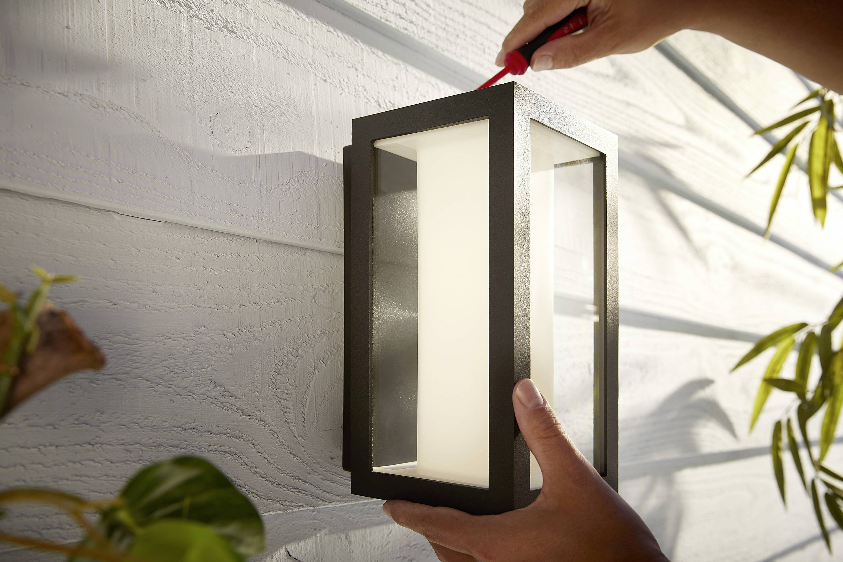 Una persona sta installando una lampada da esterno su un muro di casa alla luce del giorno, tenendo in mano un cacciavite.