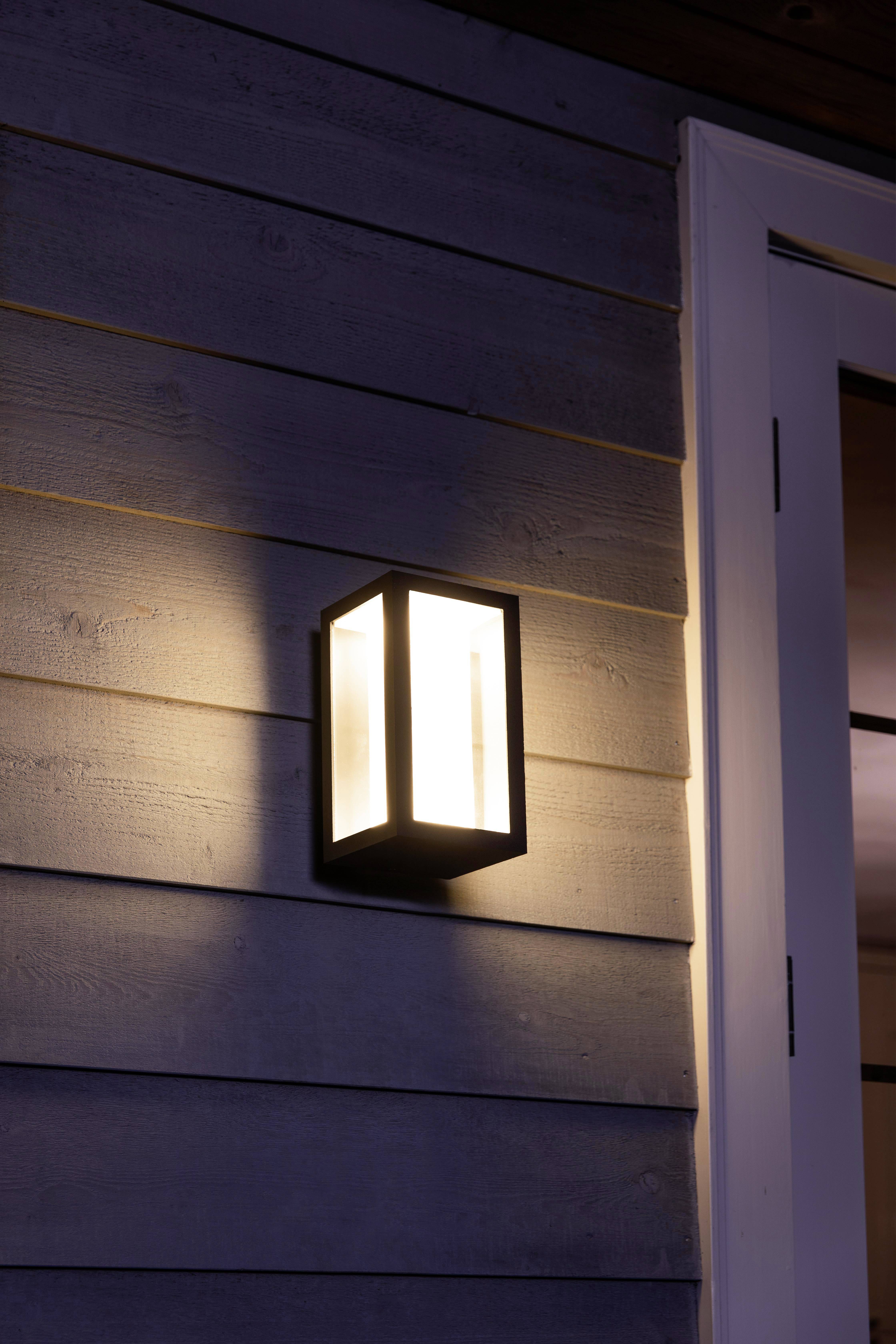 Lampada da parete esterna illumina un muro di legno. Cornice della porta a destra nell'immagine, la luce crea un'atmosfera serale calda.