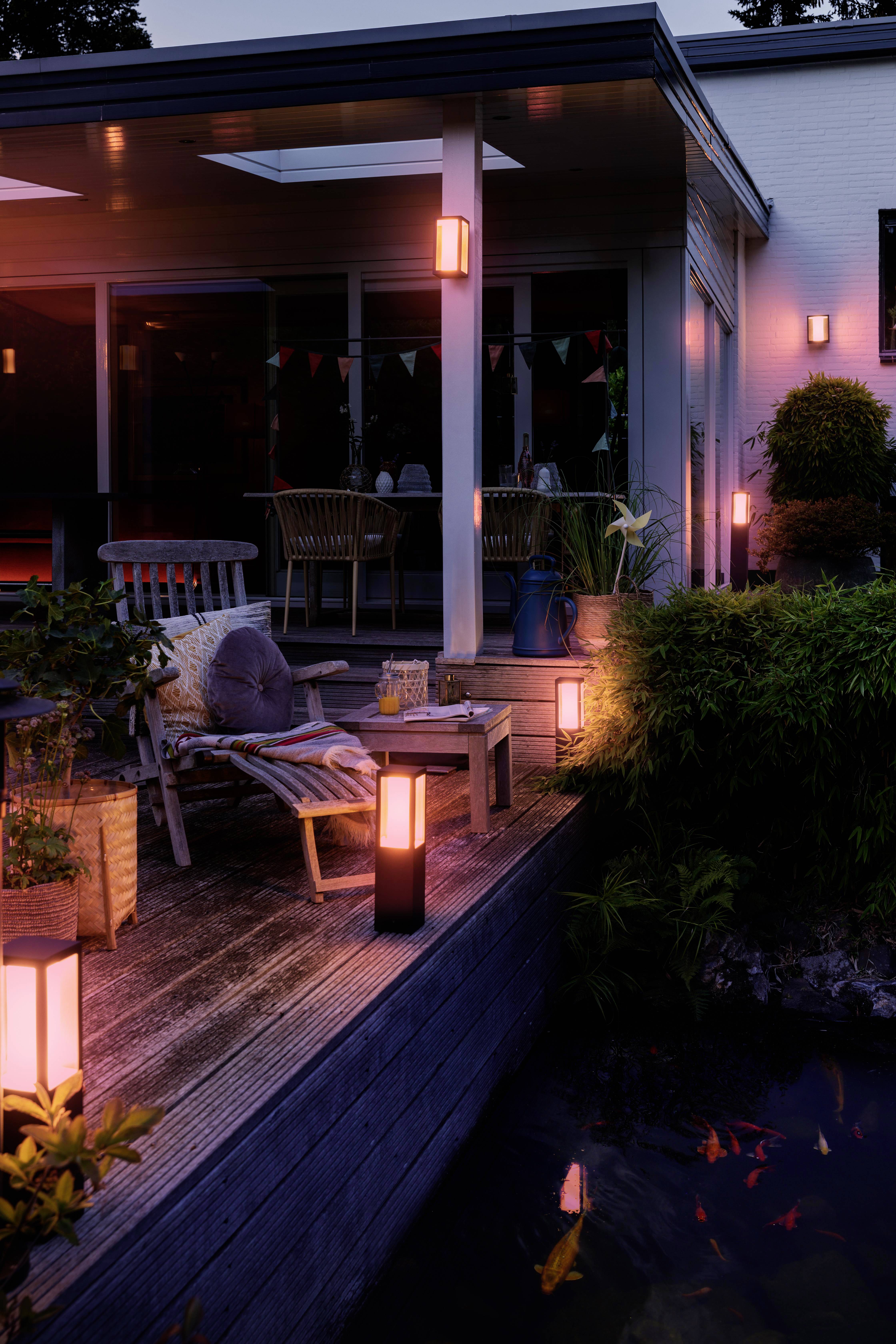 Un giardino illuminato la sera con terrazza. Lampade circondano mobili da seduta e piante vicino a uno stagno, creando un'atmosfera accogliente.
